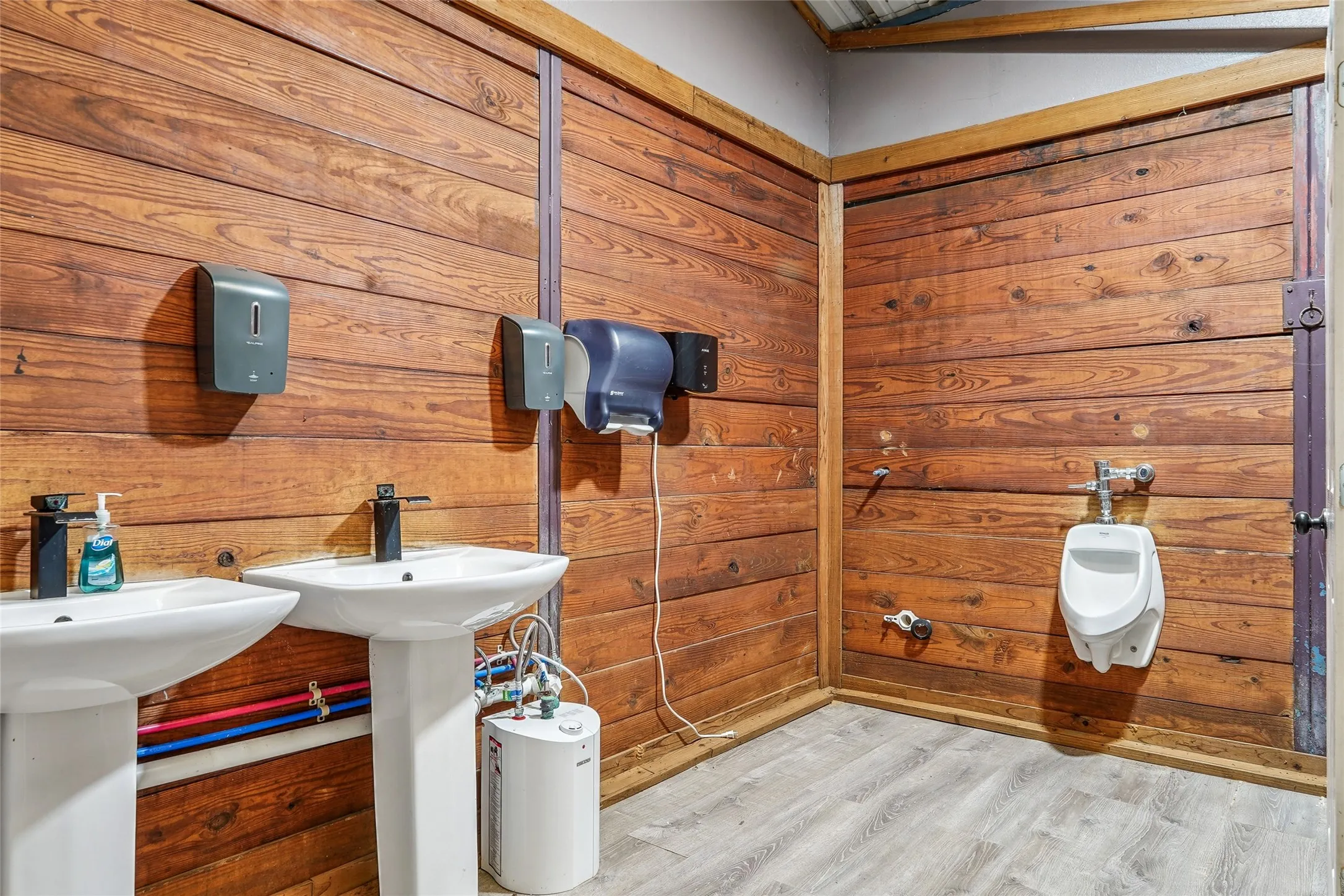 Commercial restroom featuring dual pedestal sinks with black faucets, wood-finish wall paneling, a wall-mounted urinal, and light wood-finish flooring