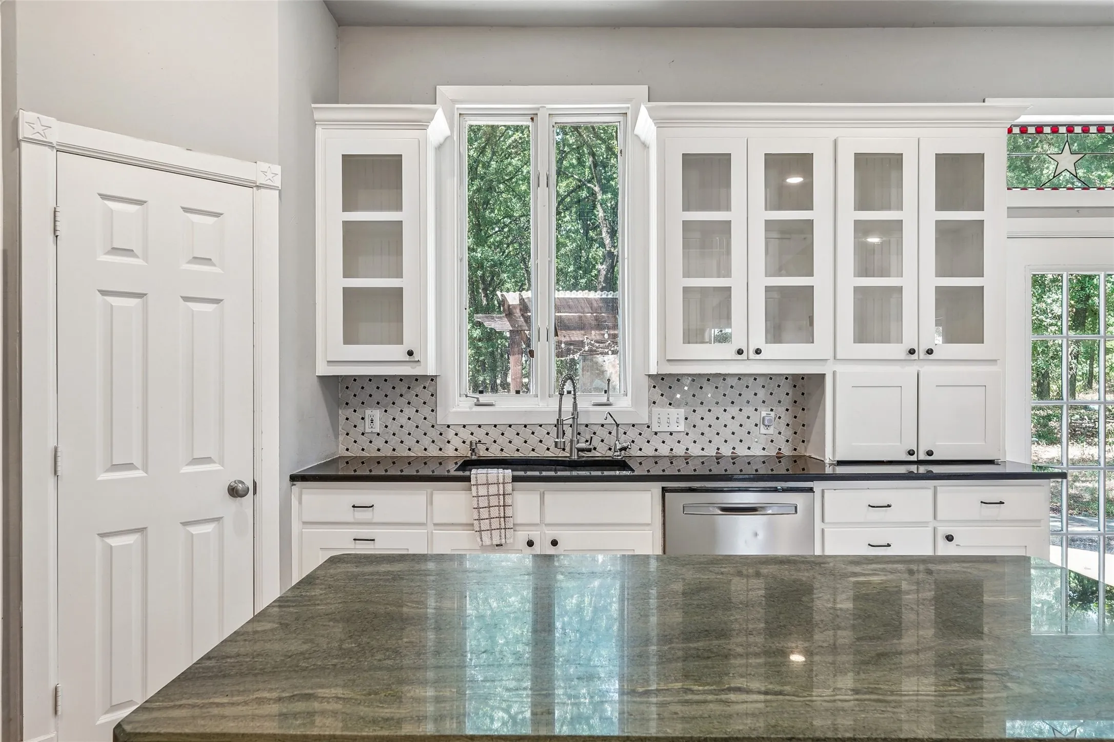 Kitchen featuring a large central island with a polished countertop, white shaker-style cabinetry with glass fronts, a black and white tiled backsplash, black countertops, and a stainless steel dishwasher