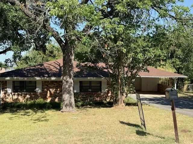 Brick exterior with a brown shingle roof, mature trees, and a covered carport