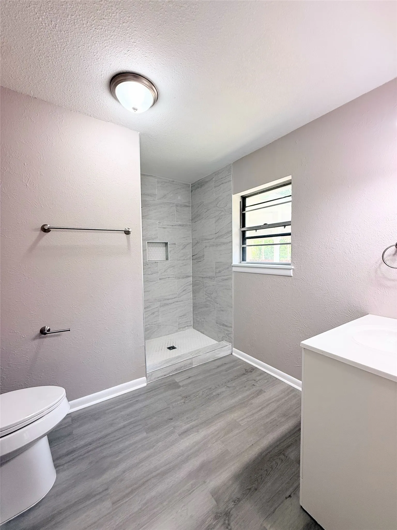 Bathroom featuring a walk-in shower with tiled surround and built-in niche, wood-finish flooring, a contemporary vanity with a white countertop, and a window for natural light