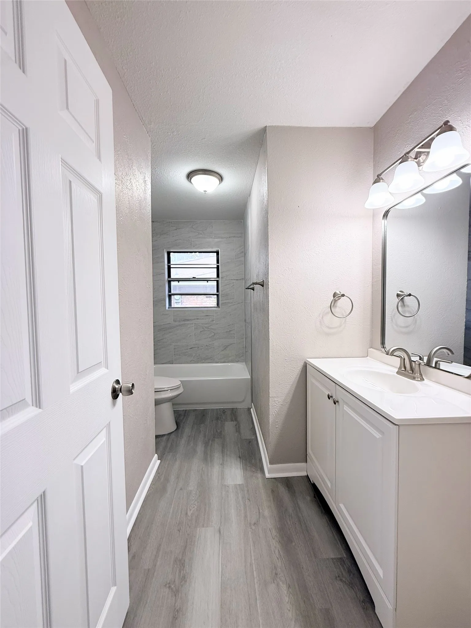 Bathroom featuring a white vanity with integrated sink, wood-finish flooring, a full bathtub with tiled surround, and a wall-mounted light fixture above the mirror