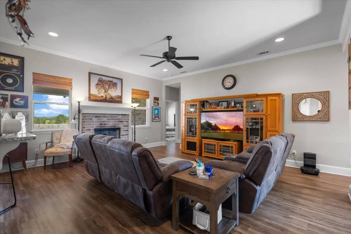 Open living area with wood-look flooring, brick fireplace, Ceiling fan and recessed lighting.