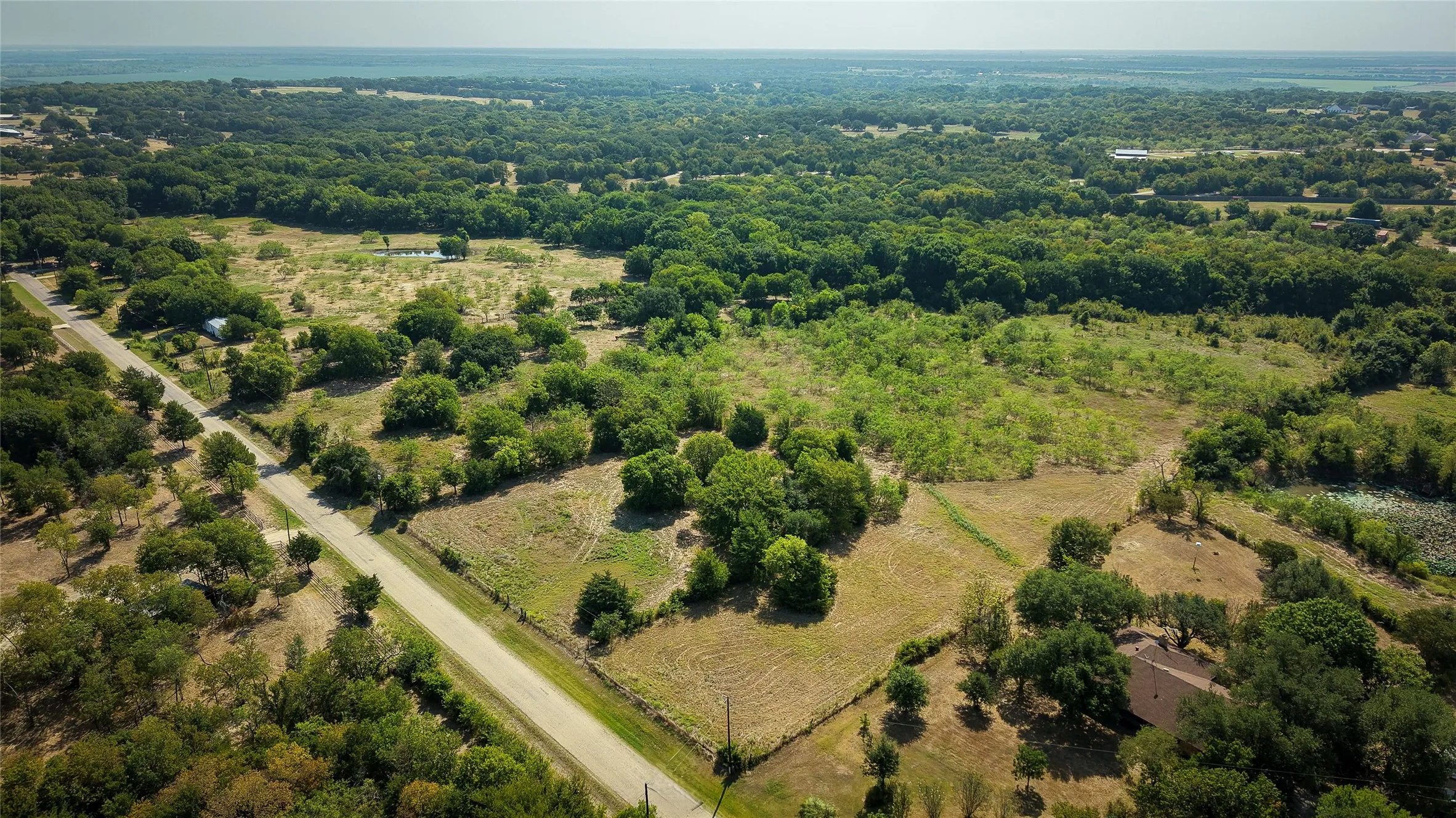 Expansive rural landscape featuring a paved road, abundant mature trees, open fields, and a distant pond