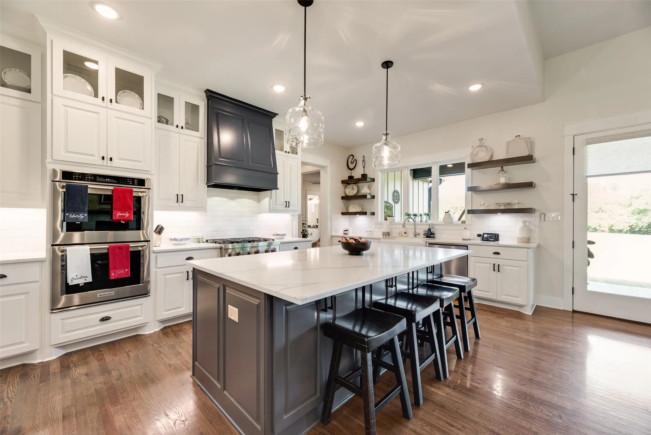 Spacious kitchen featuring a central island with countertop seating, white cabinetry, integrated stainless steel double ovens, a dark paneled range hood, and wood-finish flooring