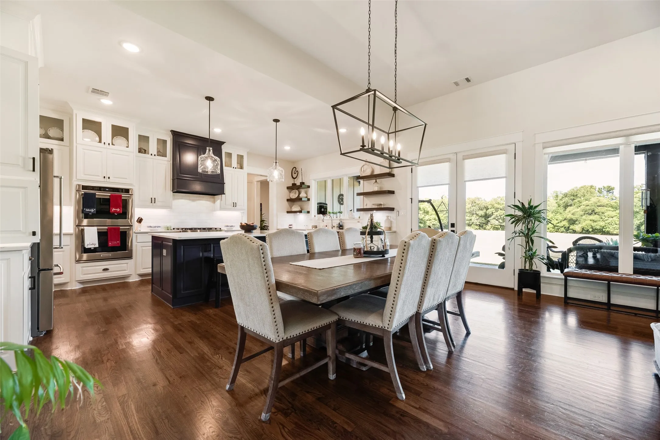 Open-concept living space featuring rich wood-finish flooring, a contemporary dining area with a linear chandelier, a kitchen with white cabinetry and a contrasting dark island, stainless steel double ovens, and large windows bringing in natural light