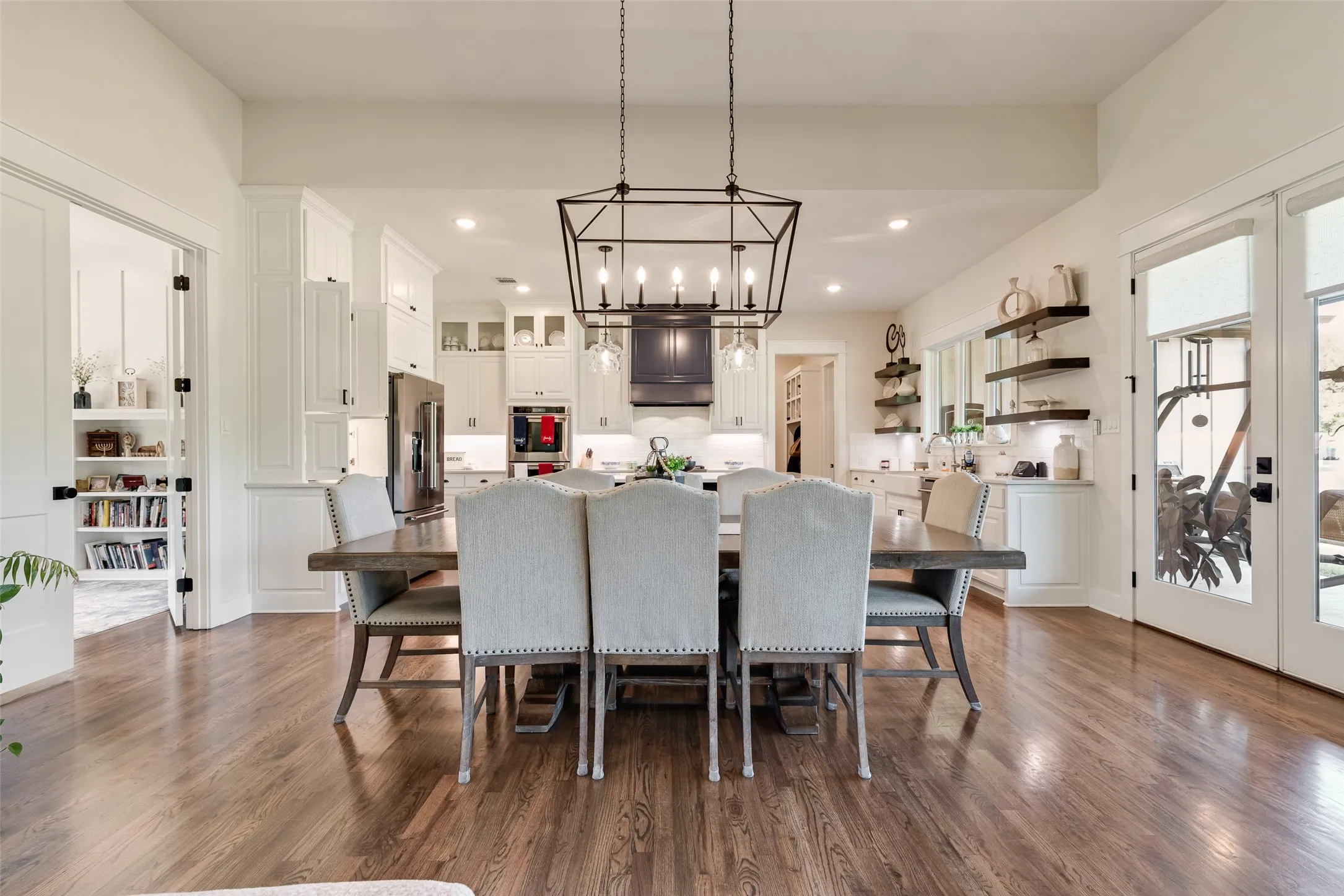 Expansive open-concept living area featuring rich wood-finish flooring, a contemporary black geometric chandelier, white cabinetry, integrated stainless steel appliances, and French doors