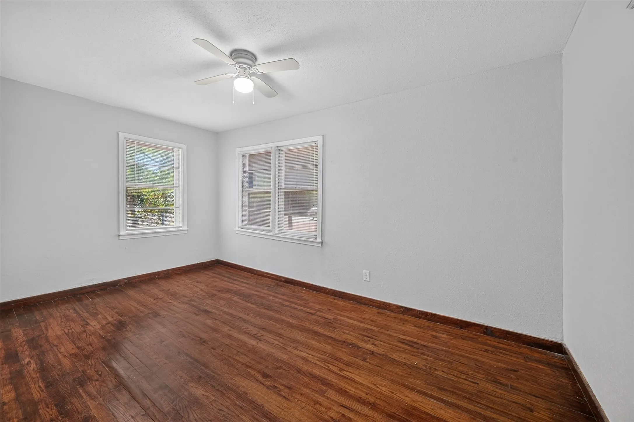 Wood-finish flooring, white walls, two windows, and a ceiling fan with integrated lighting