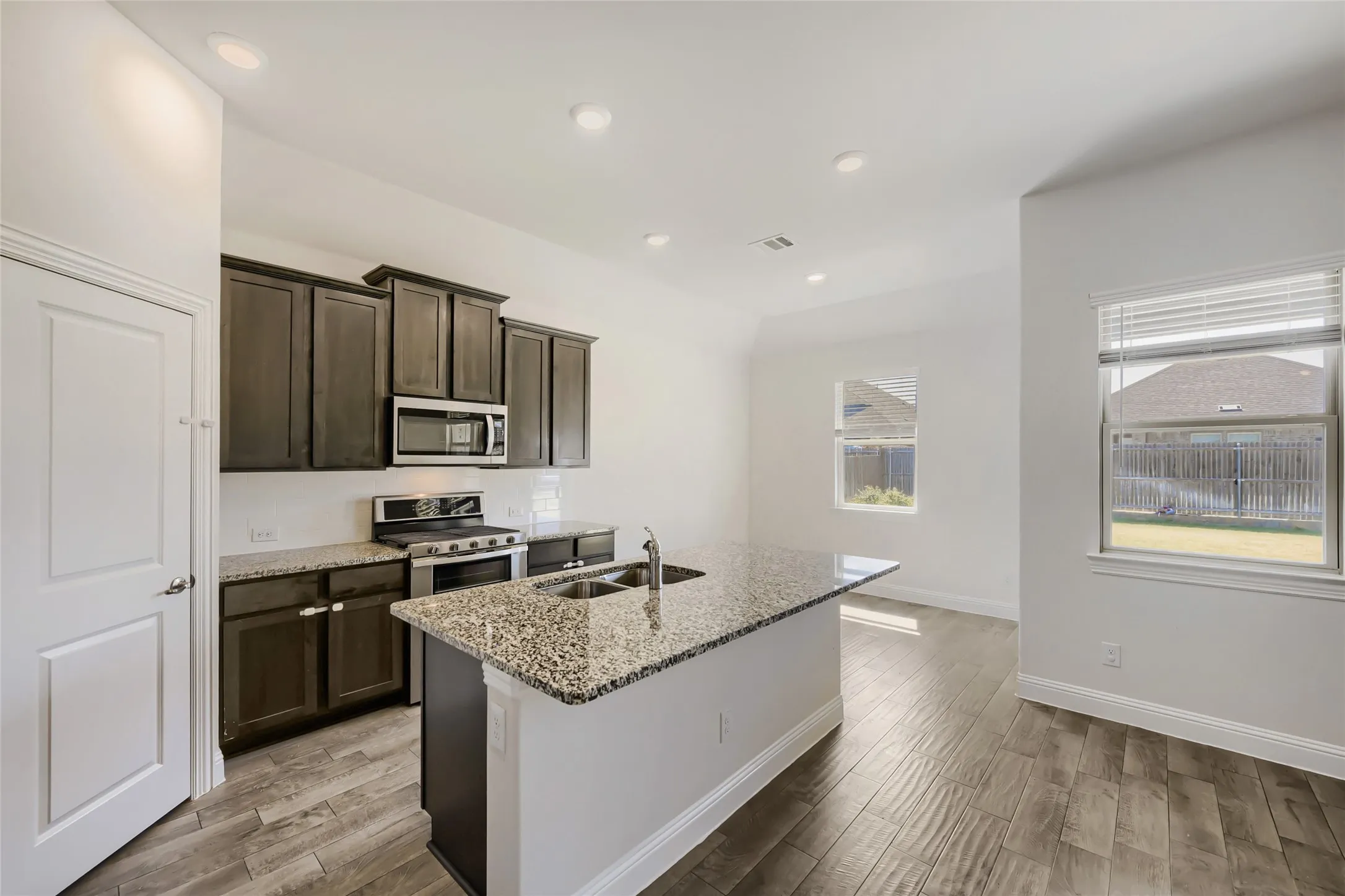 Open-concept kitchen featuring an island with an integrated double sink, granite countertops, dark wood cabinetry, and stainless steel appliances