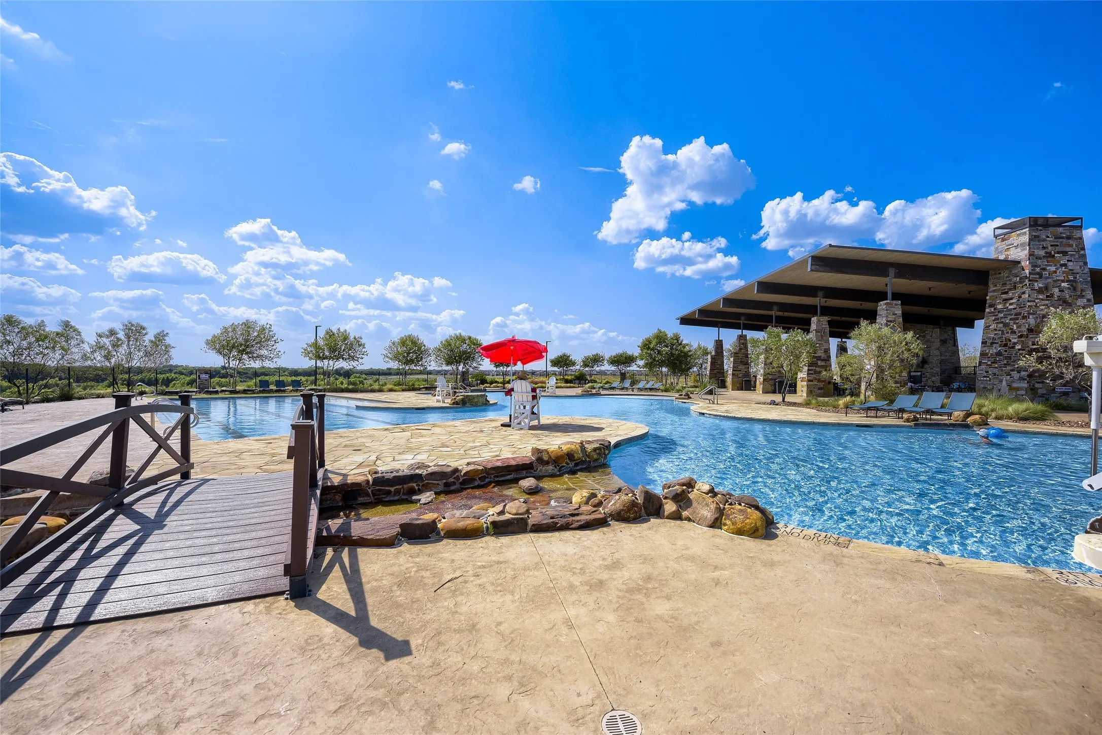 Expansive outdoor aquatic area featuring a multi-level pool, a decorative bridge with dark wood-finish decking, and a covered lounge structure with stone-clad columns