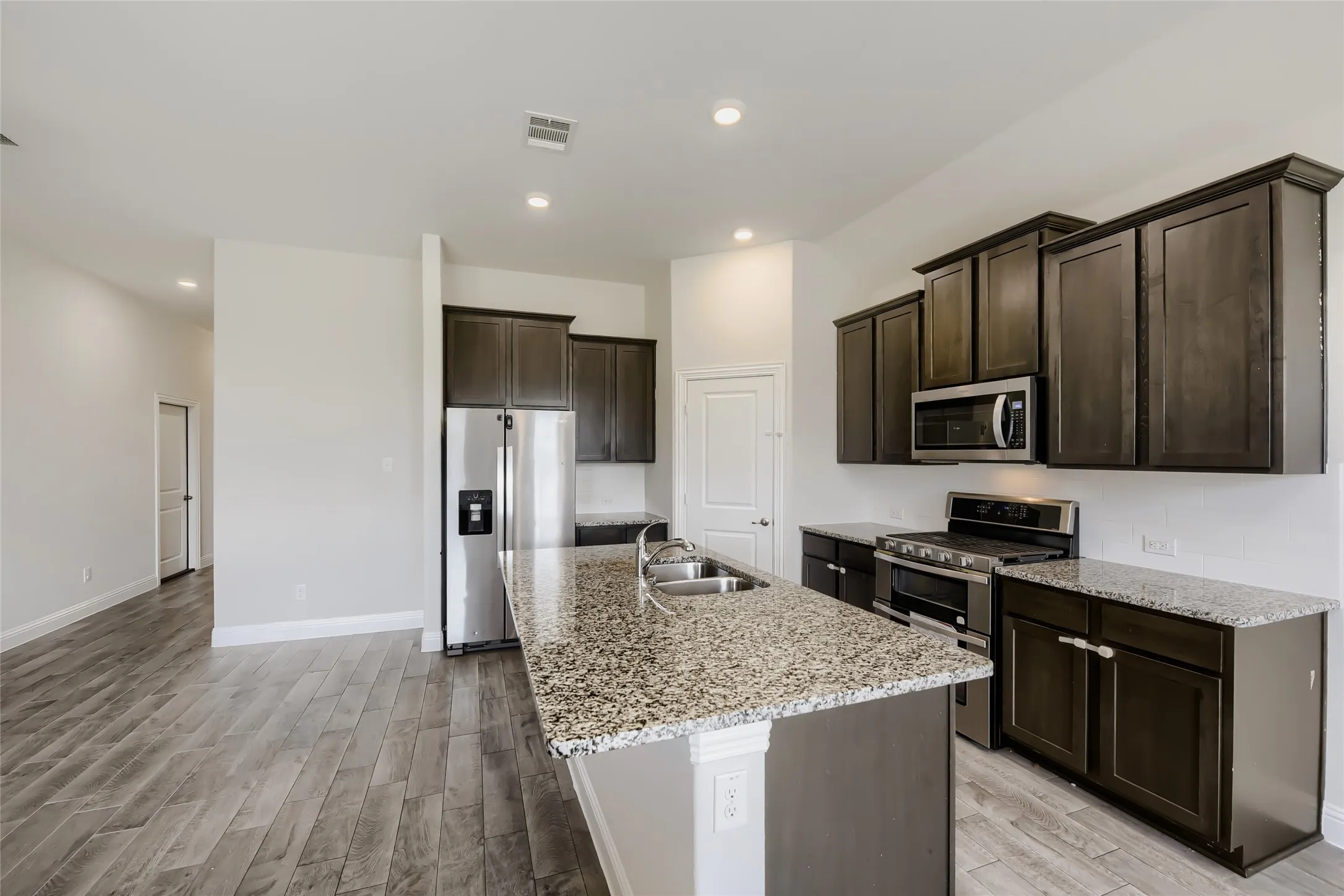 Kitchen featuring wood-finish flooring, dark wood cabinetry, stainless steel appliances, speckled countertops, and an island with an undermount double-basin sink