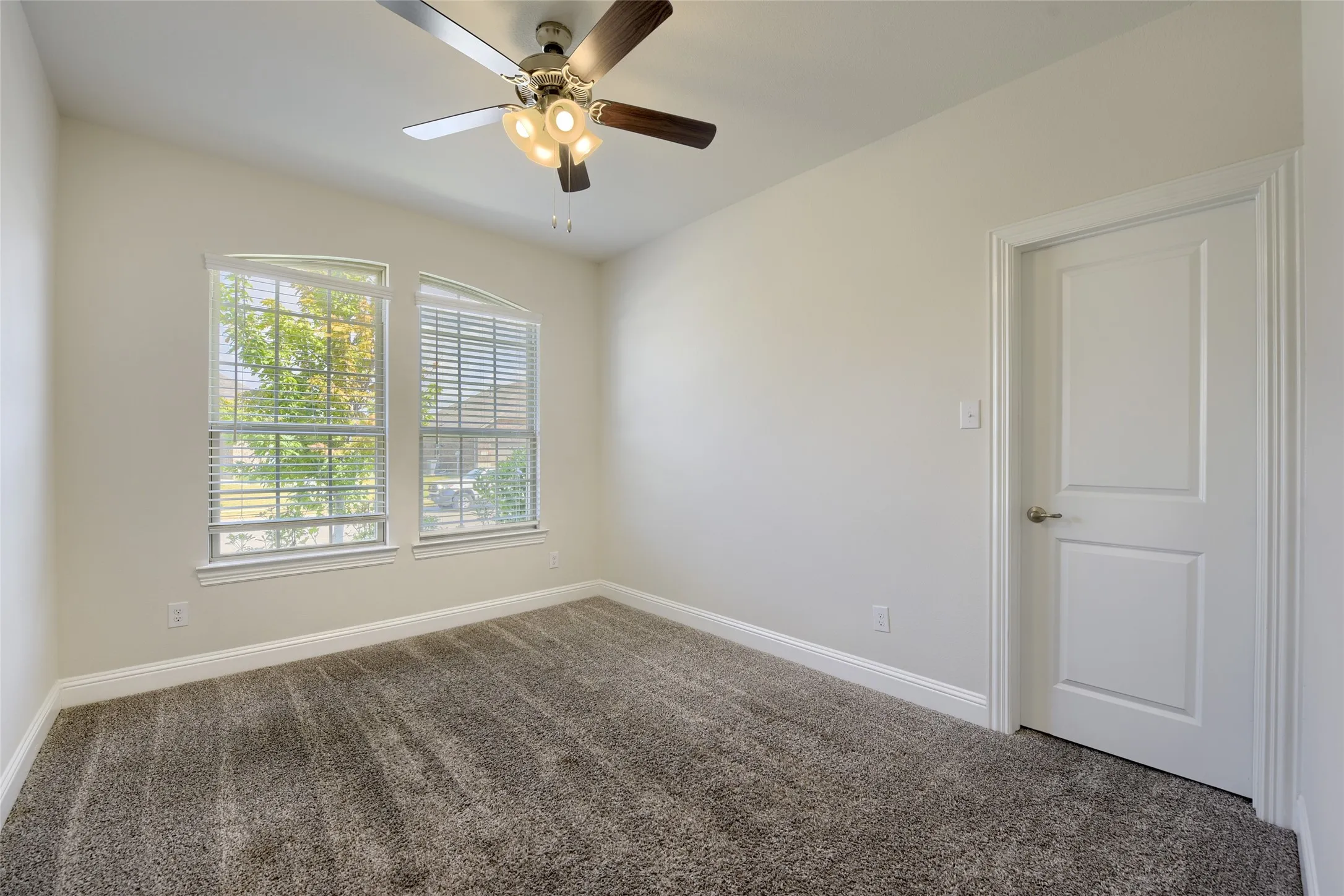 Neutral-toned bedroom featuring twin arched windows with blinds, warm gray carpeting, and a ceiling fan with integrated lighting