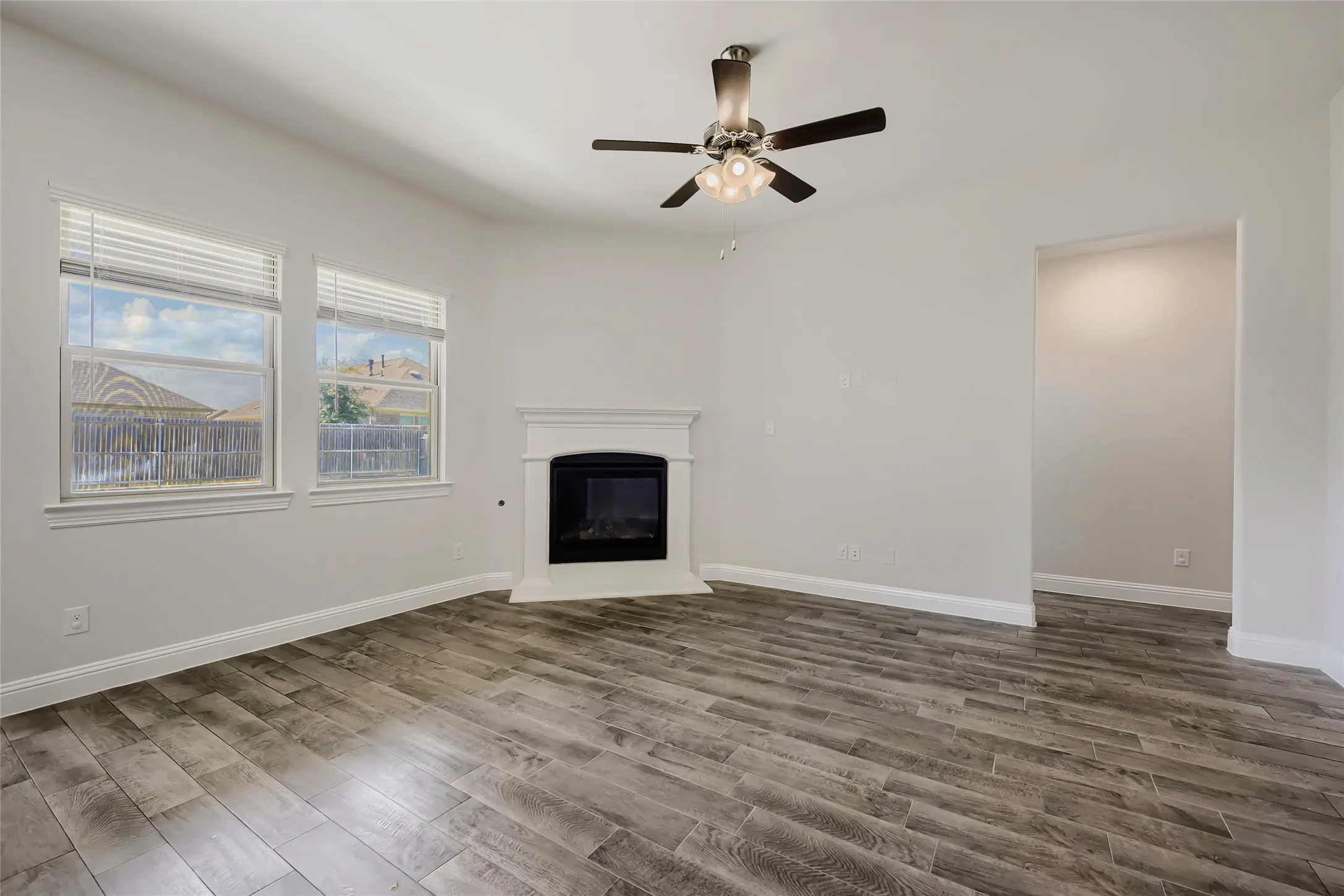 Spacious room featuring wood-finish flooring, a corner-set fireplace with white mantel, two windows with blinds, a ceiling fan with integrated lighting, and a wide entryway