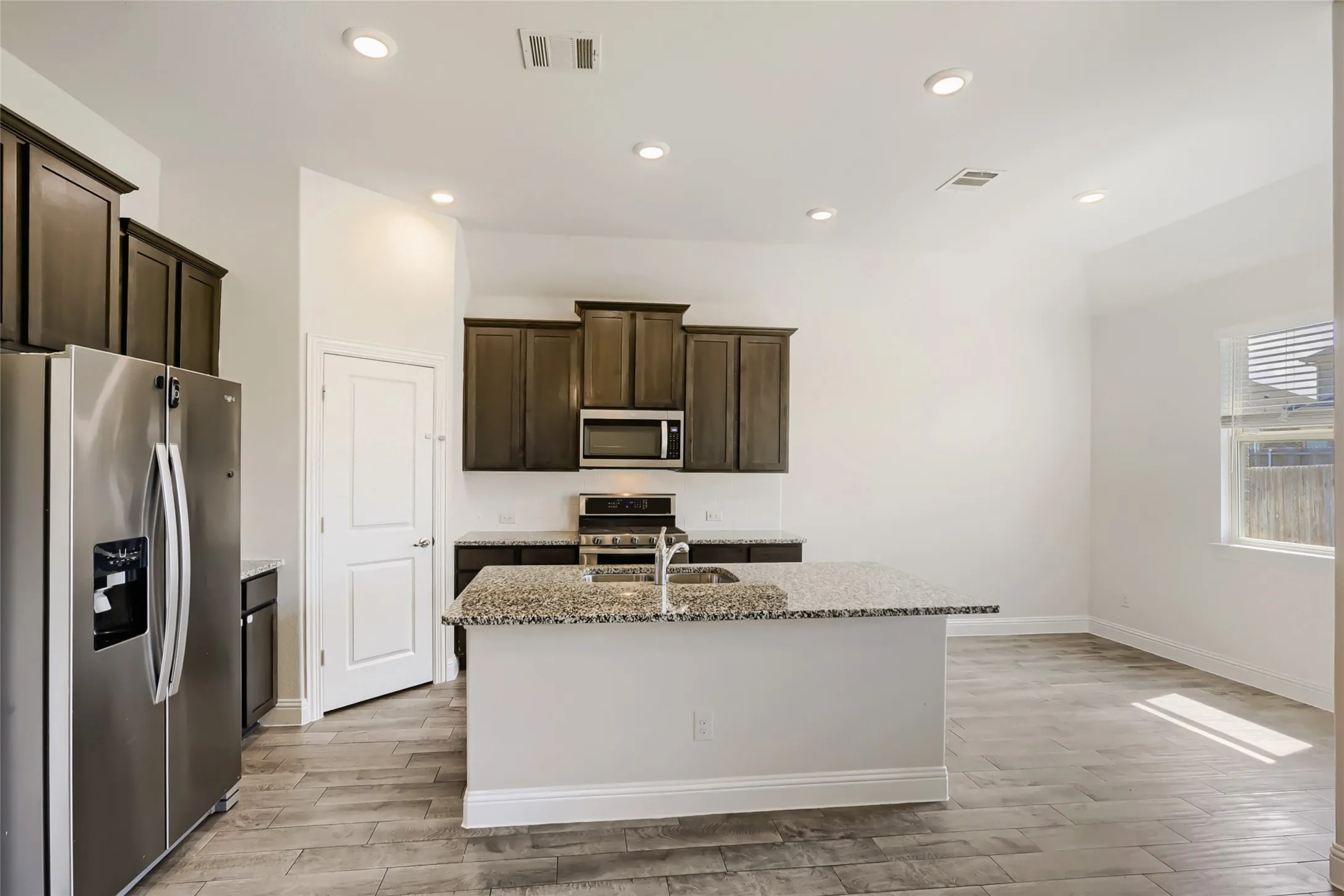 Open-concept kitchen featuring an island with a built-in sink, stainless steel appliances, dark wood cabinetry, light-toned countertops, and wood-finish flooring