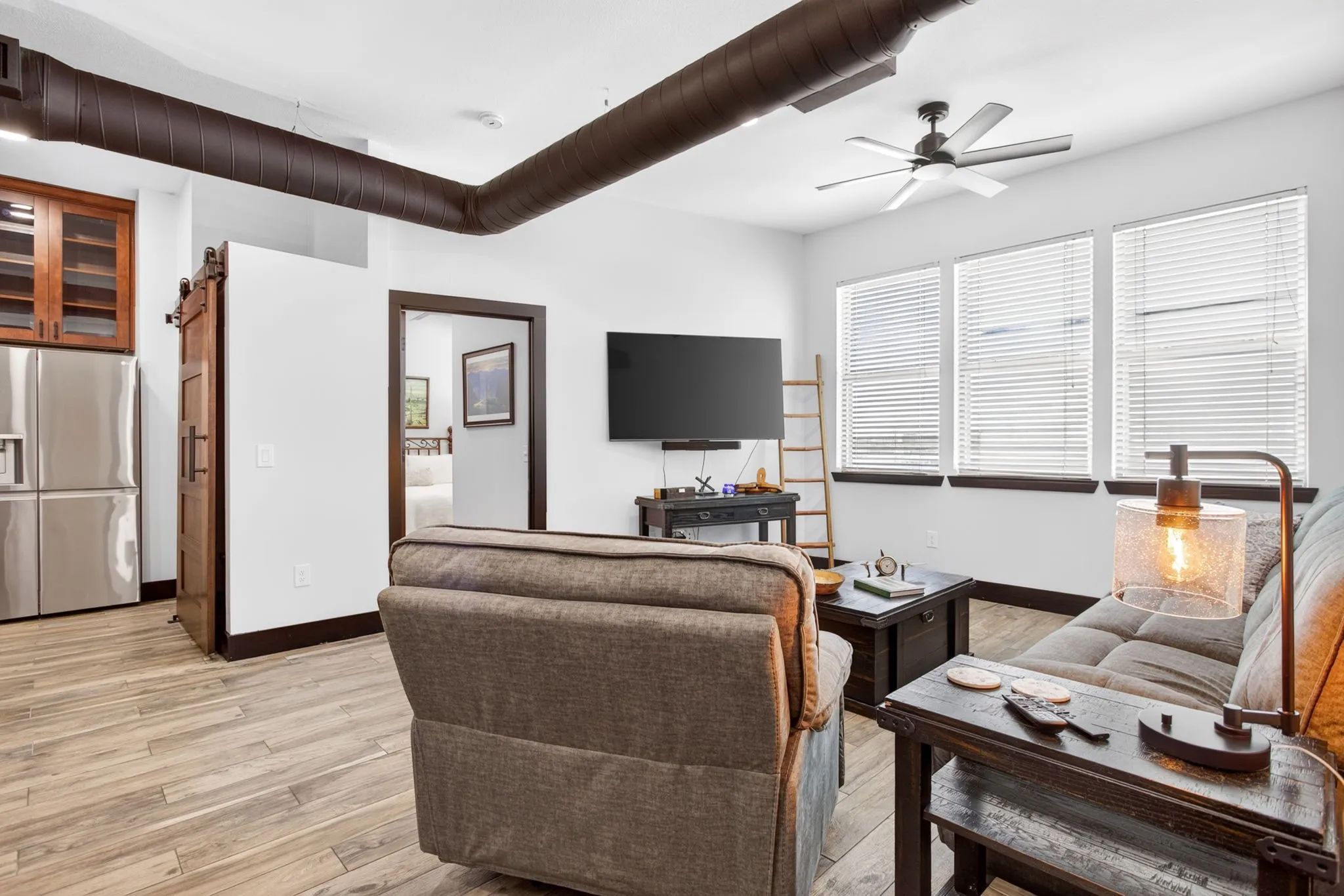 Open-concept living space featuring wood-finish flooring, a kitchen with stainless steel appliances and wood cabinetry, and an exposed brick accent wall