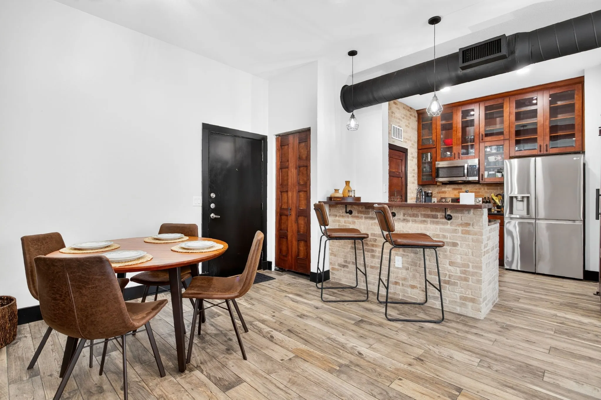 Open-concept living space featuring wood-finish flooring, a kitchen with stainless steel appliances and wood cabinetry, and an exposed brick accent wall