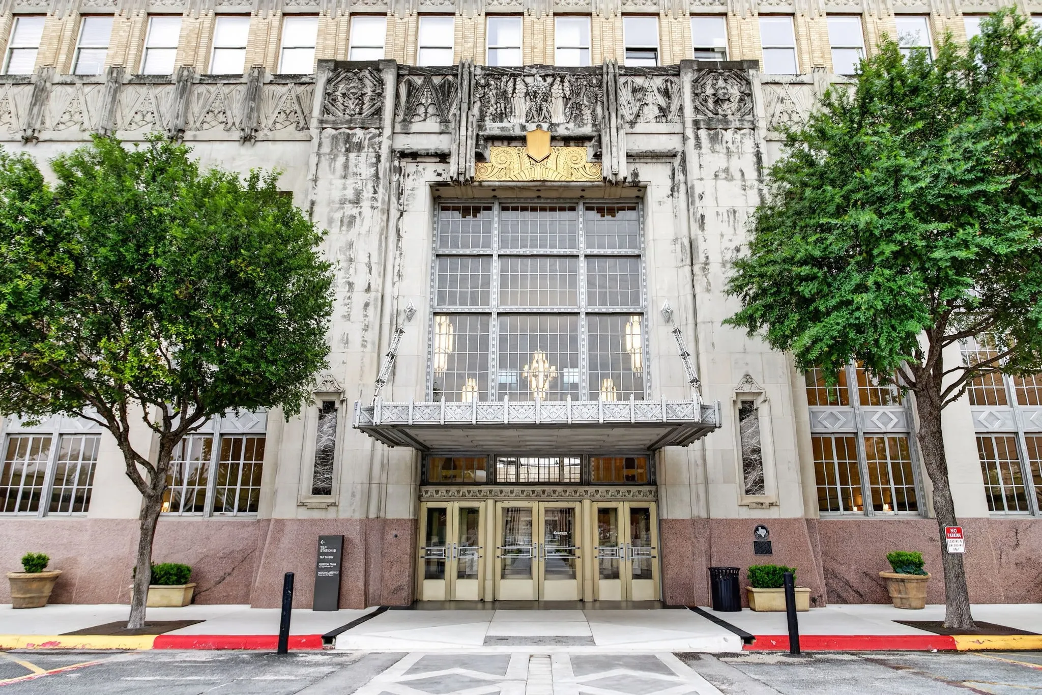 Building exterior featuring an ornate entrance, decorative facade with sculptural elements, large multi-pane windows, a detailed metal awning, and mature trees framing the entry