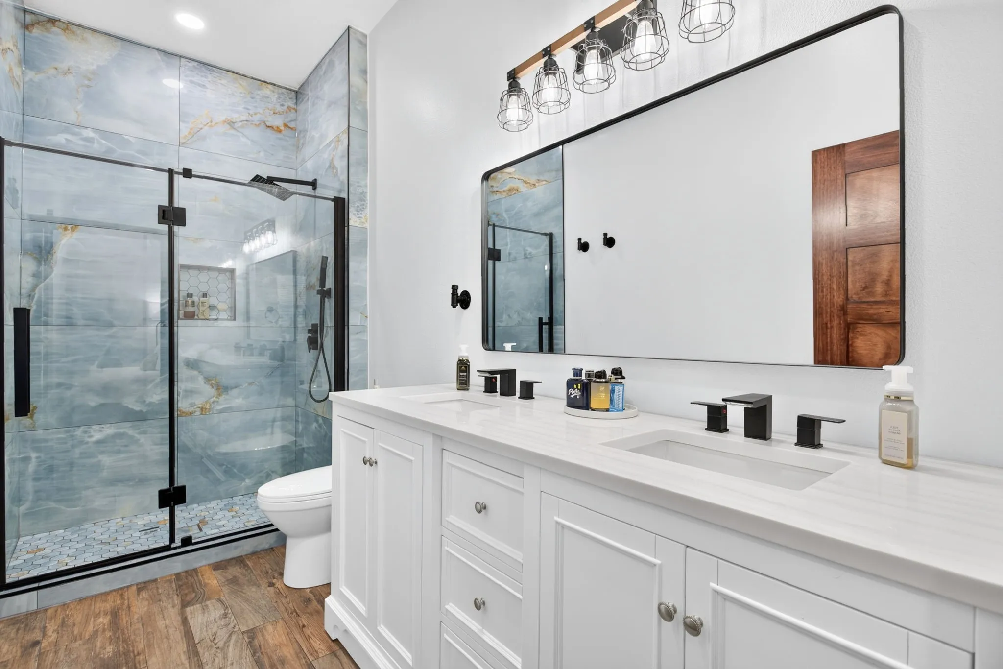 Bathroom featuring a dual vanity with white cabinetry and integrated sinks, a wide rectangular mirror, and a walk-in shower with blue-toned tile and a glass enclosure