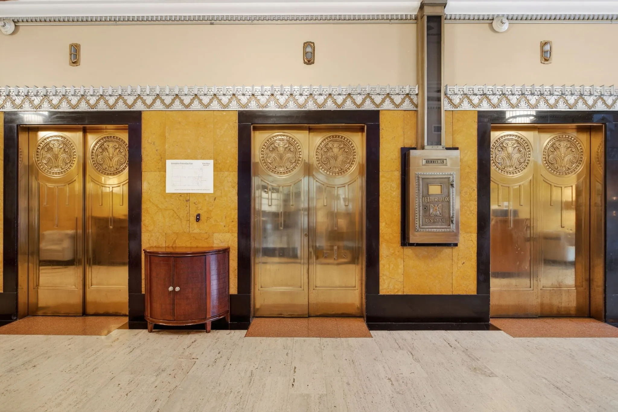 Grand lobby featuring three ornate golden elevator doors with intricate circular medallions, set against a two-tone wall with dark trim and a light, textured upper section