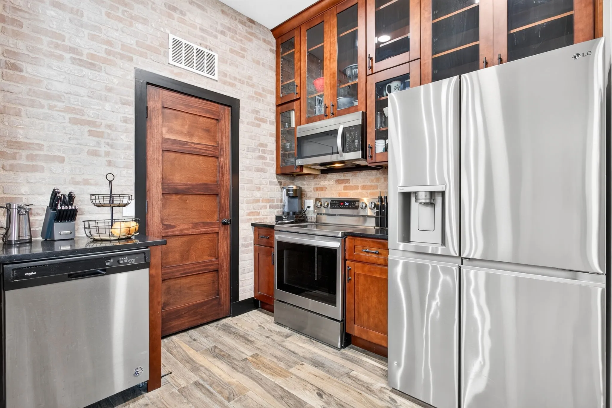 Kitchen featuring light brick wall, wood-finish flooring, stainless steel appliances, dark wood cabinetry with glass fronts, and dark countertops