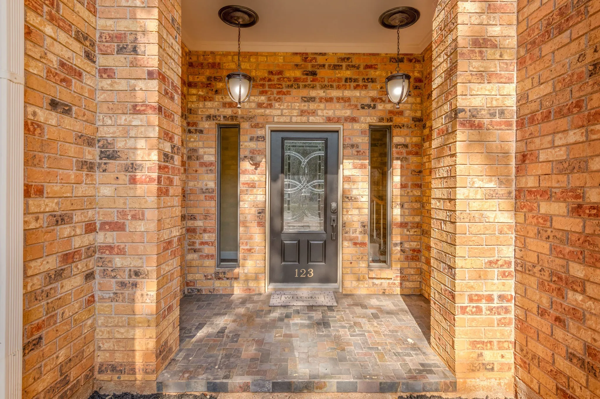 Welcoming covered porch surrounded with brick and accented with stunning hanging fixtures, black decorative glass door and side windows.