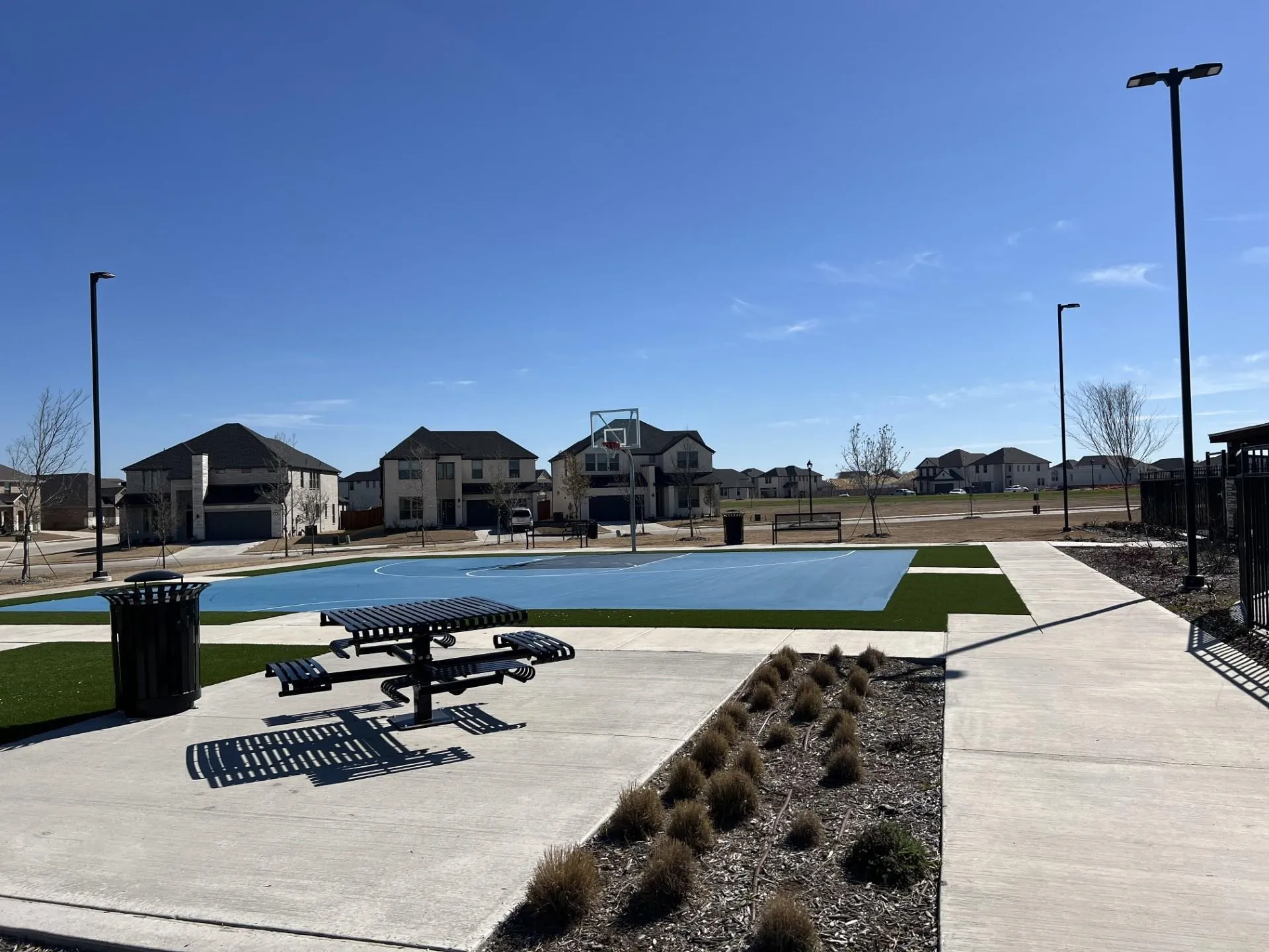 Community recreational area featuring a blue-surfaced basketball court, a picnic table with seating, concrete pathways, and integrated street lighting
