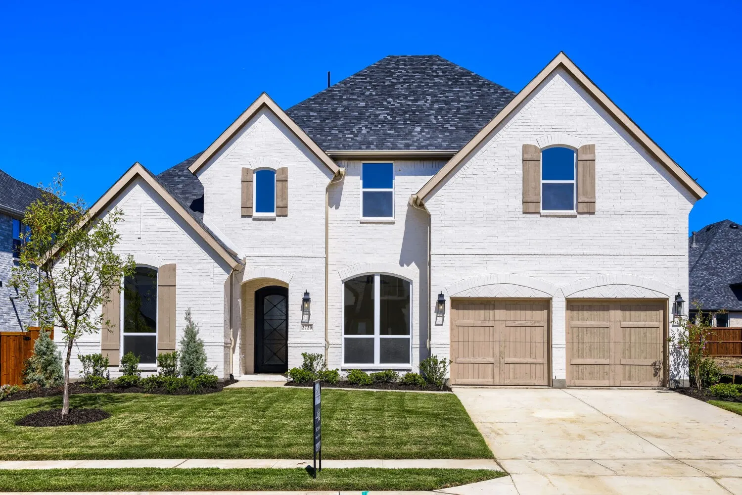 White brick exterior with a dark asphalt shingle roof, arched entryway, wood-finish garage doors, and decorative window shutters