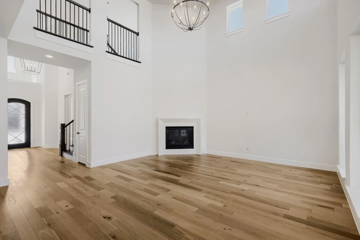 Double-height ceiling, wood-finish flooring, white fireplace with mantle, decorative chandelier, and black wrought iron stair railing