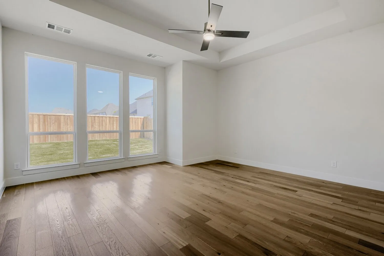 Tray ceiling with a modern ceiling fan, wood-finish flooring, and multiple large windows overlooking a fenced yard
