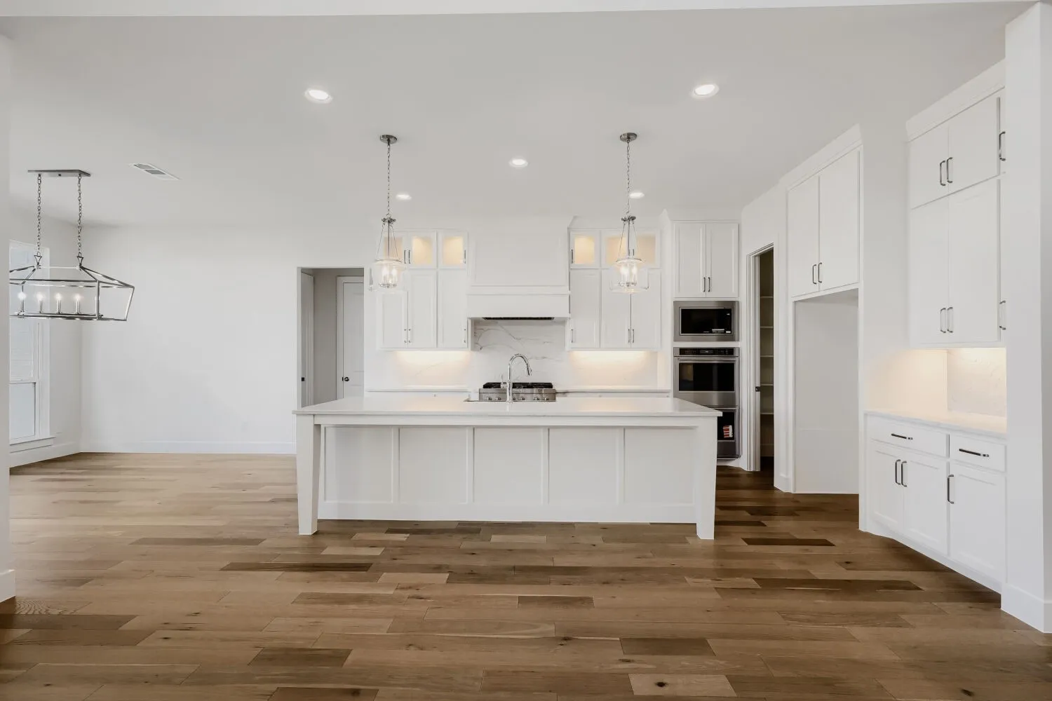 Kitchen featuring a large island with sink, stainless steel built-in appliances, white shaker-style cabinetry, pendant lighting, and wood-finish flooring