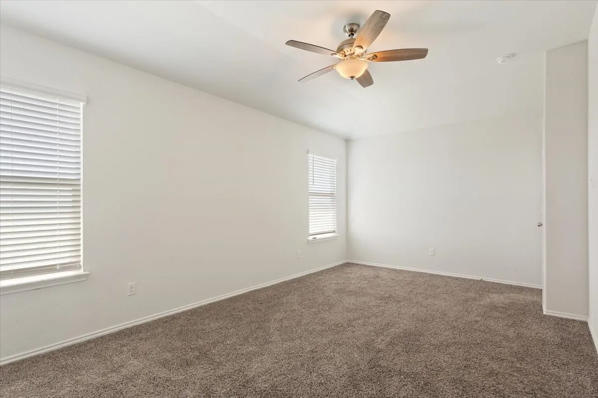 Expansive room featuring neutral-toned carpet flooring, two windows with blinds, a ceiling fan with integrated lighting, and white baseboards