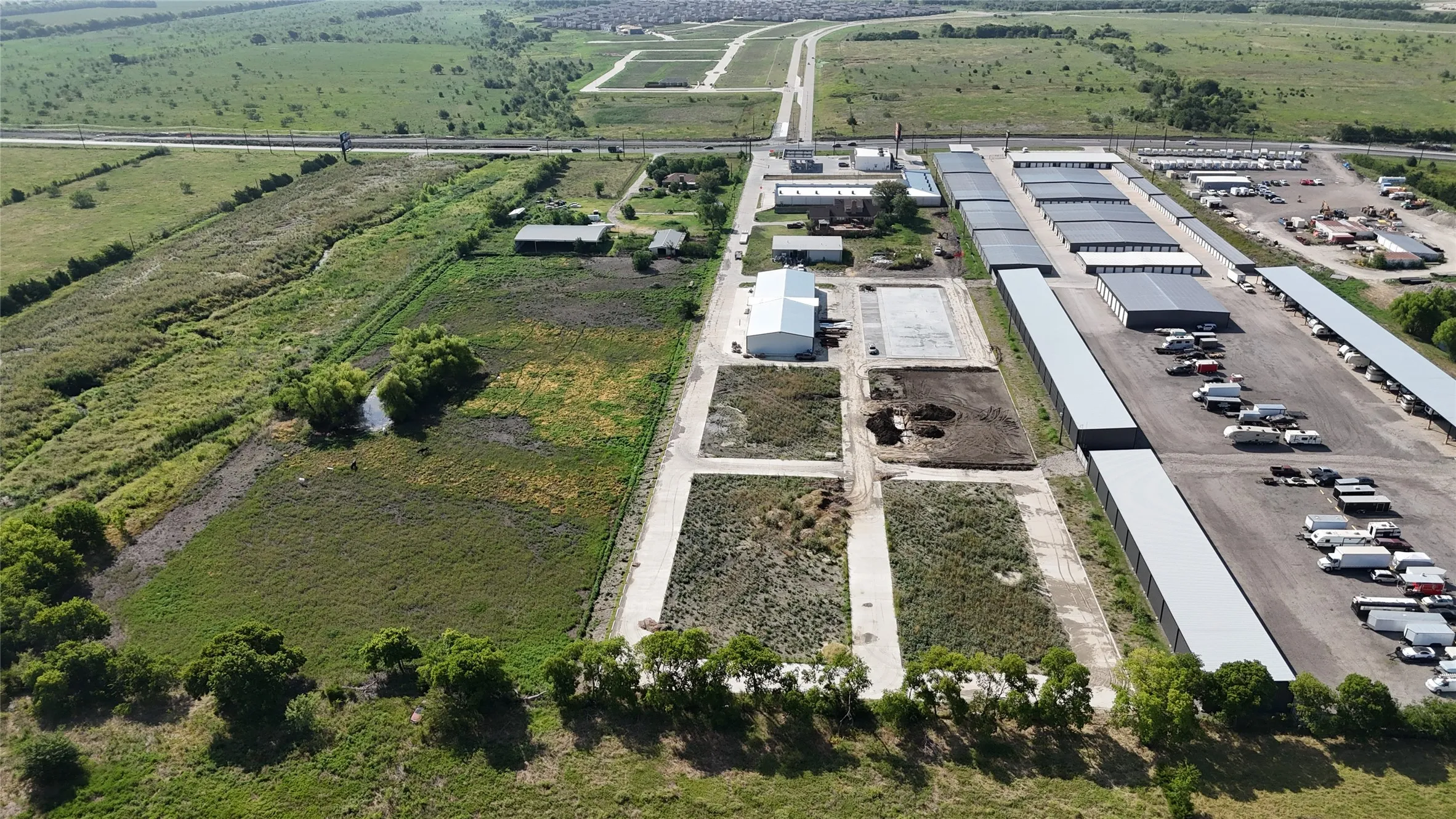 Expansive property featuring multiple large industrial buildings with metal roofs, extensive paved areas for vehicle storage, and undeveloped land