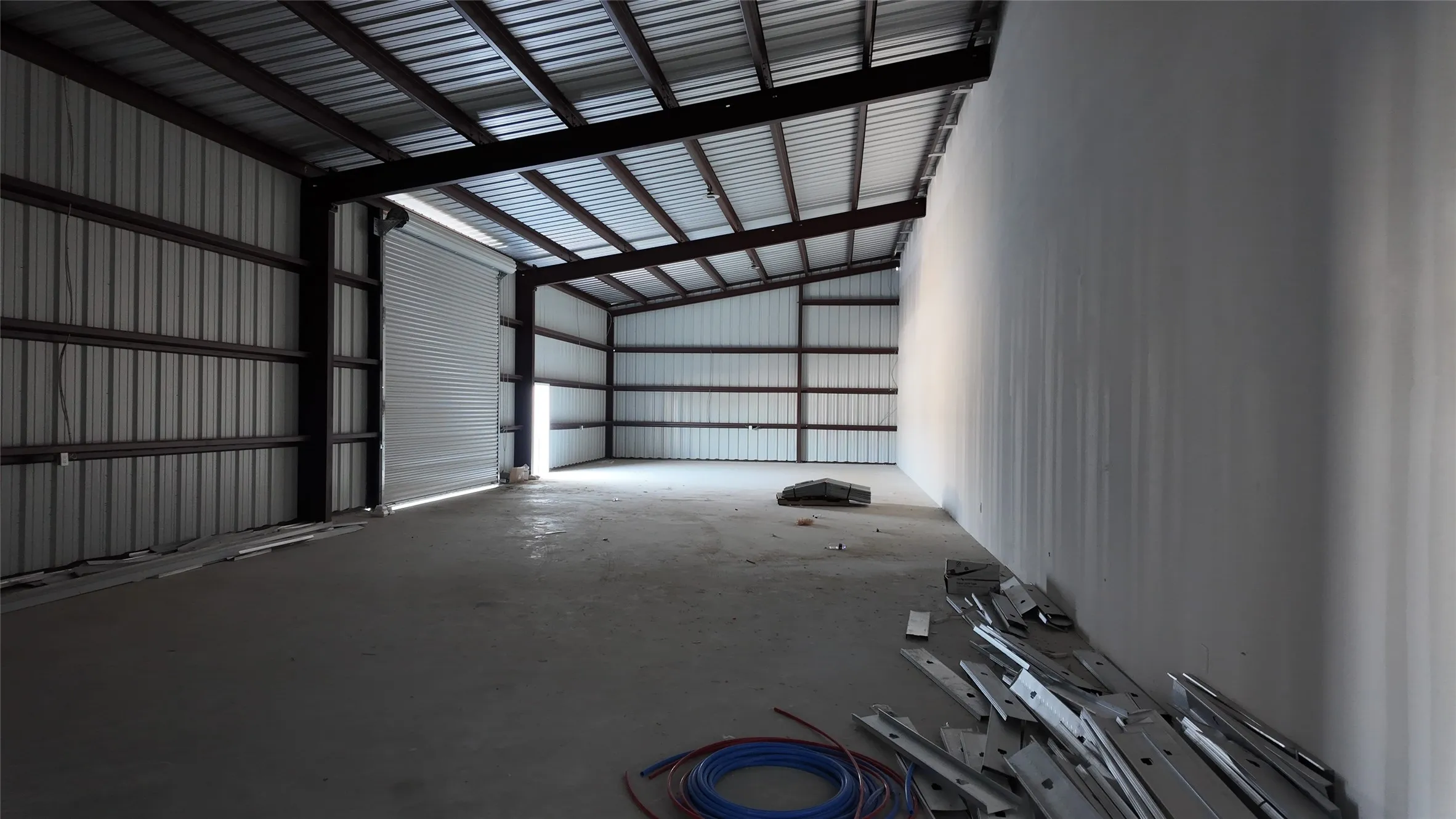 Spacious interior featuring a concrete floor, high corrugated metal ceilings, exposed metal beams, a large roll-up door, and a partially finished drywall wall