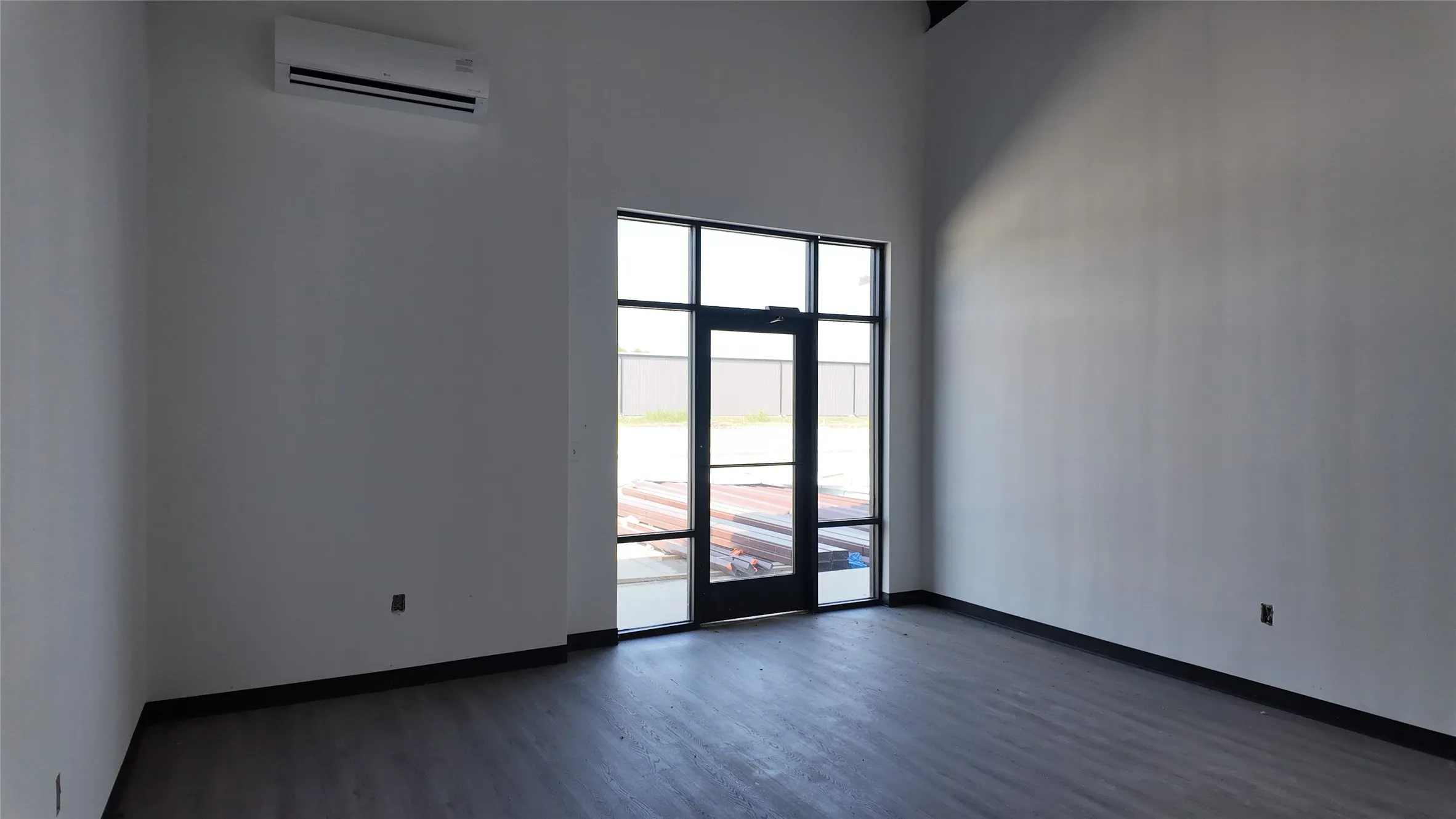 Bright, open interior space featuring expansive glass entry doors with black-frame detailing, light-filled walls, and dark wood-finish flooring throughout