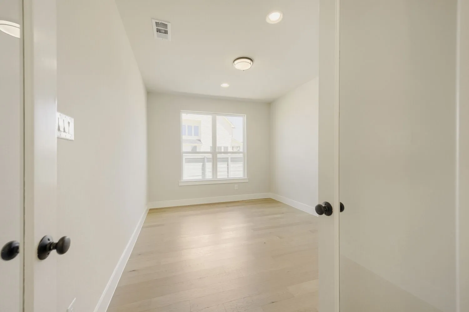 Spacious room featuring light wood-finish flooring, recessed lighting, a central flush-mount ceiling light, and a large window with white trim