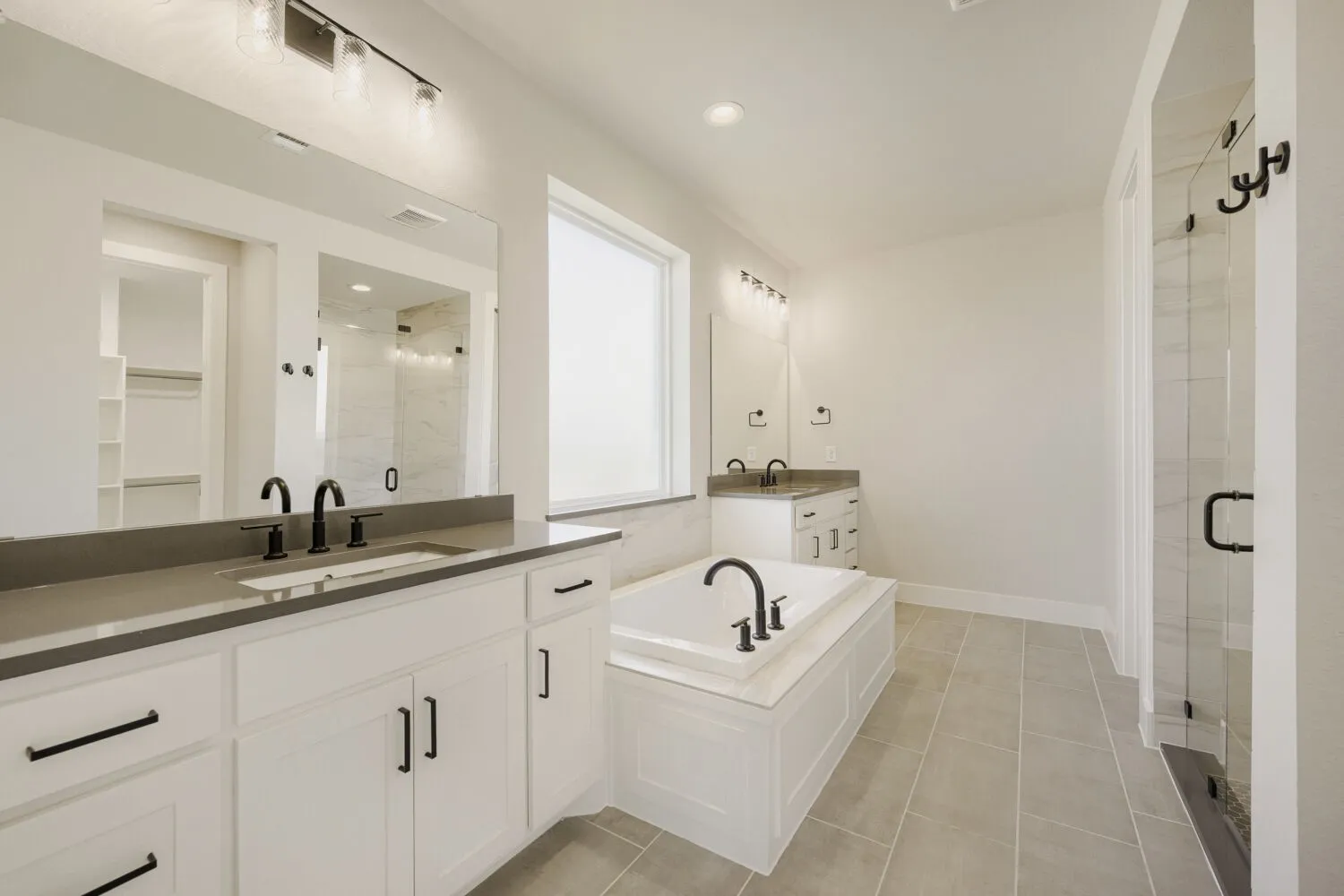 Contemporary main bathroom featuring dual white vanities with dark countertops, matte black hardware, a built-in soaking tub, a frameless glass shower, and light-toned tile flooring