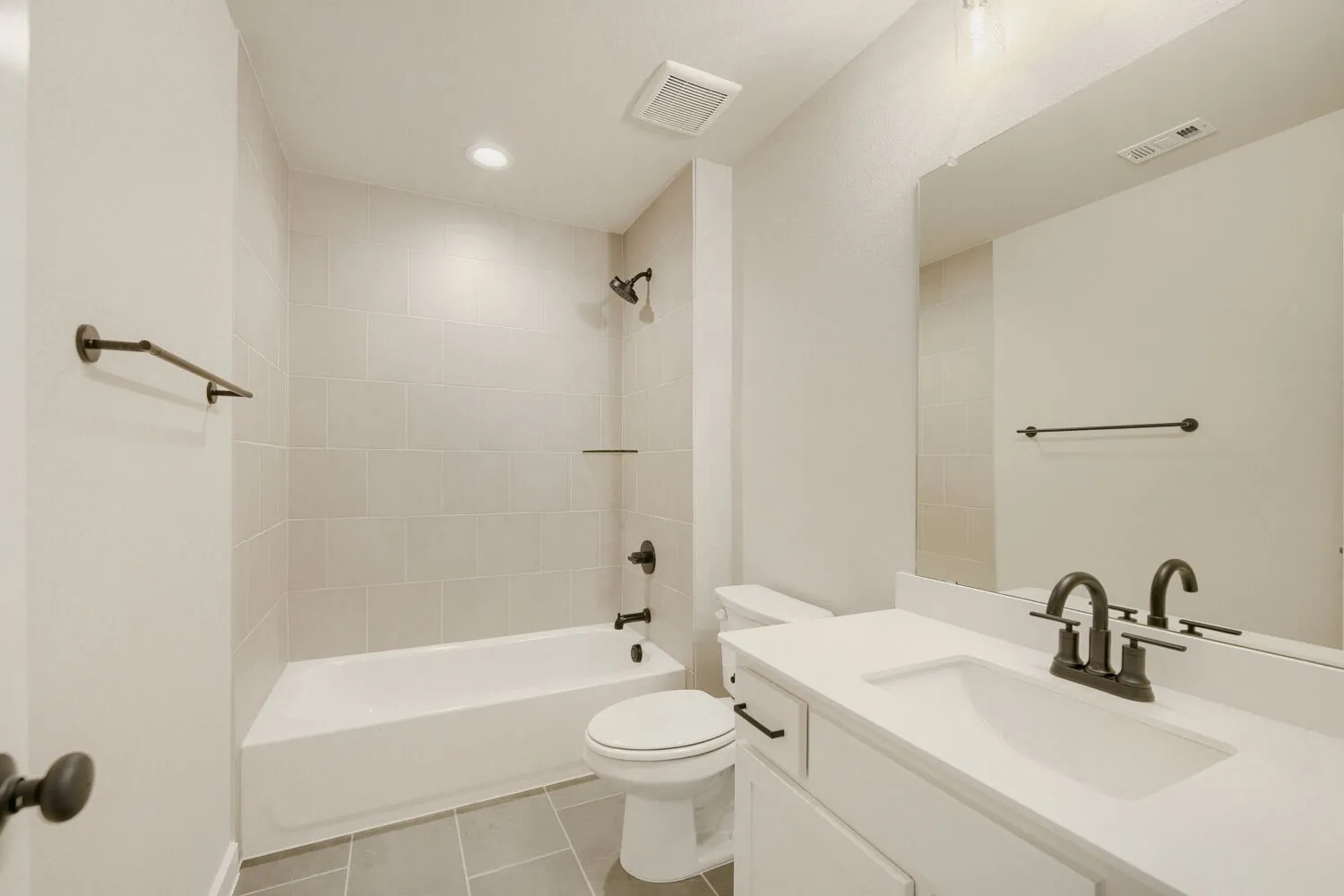Contemporary bathroom featuring a white bathtub with tile surround, dark bronze fixtures, a single vanity with a white countertop and integrated sink, and gray floor tiles