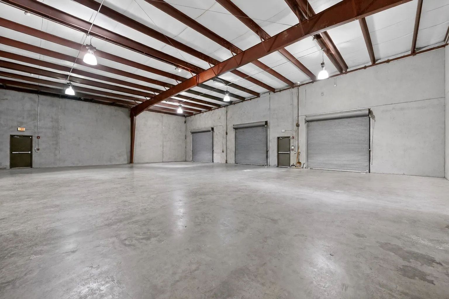 Spacious industrial interior featuring a concrete floor, concrete walls, multiple roll-up doors, and exposed metal beam ceiling with integrated lighting