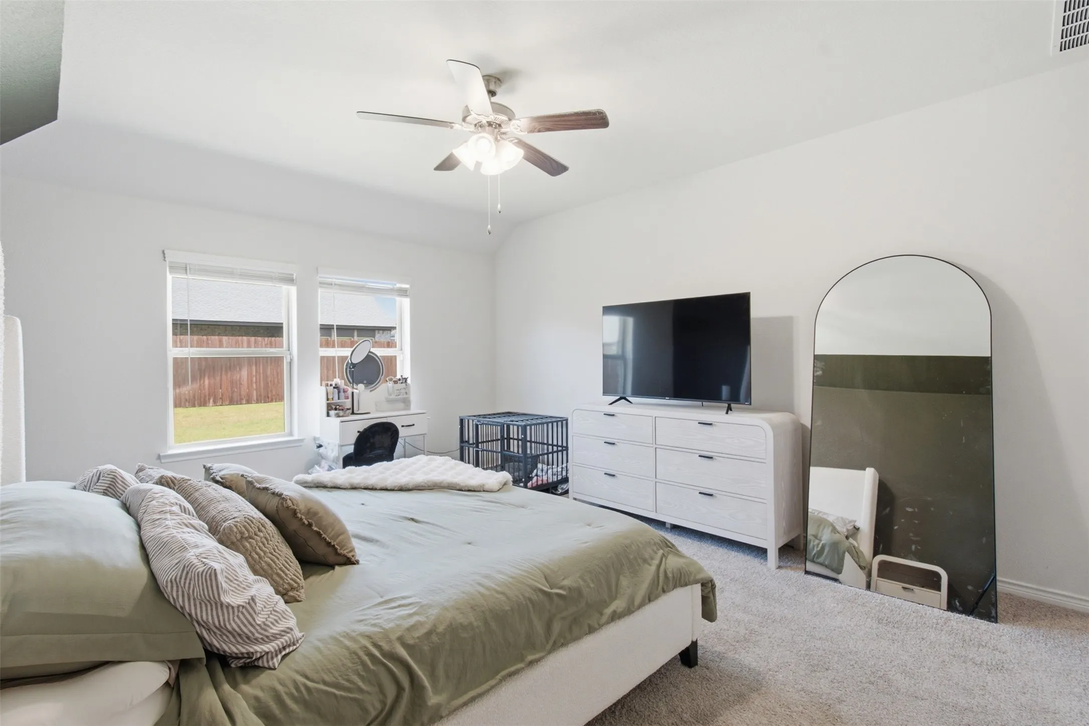 Carpeted bedroom featuring white walls, a ceiling fan with light fixture, and two windows overlooking a fenced backyard