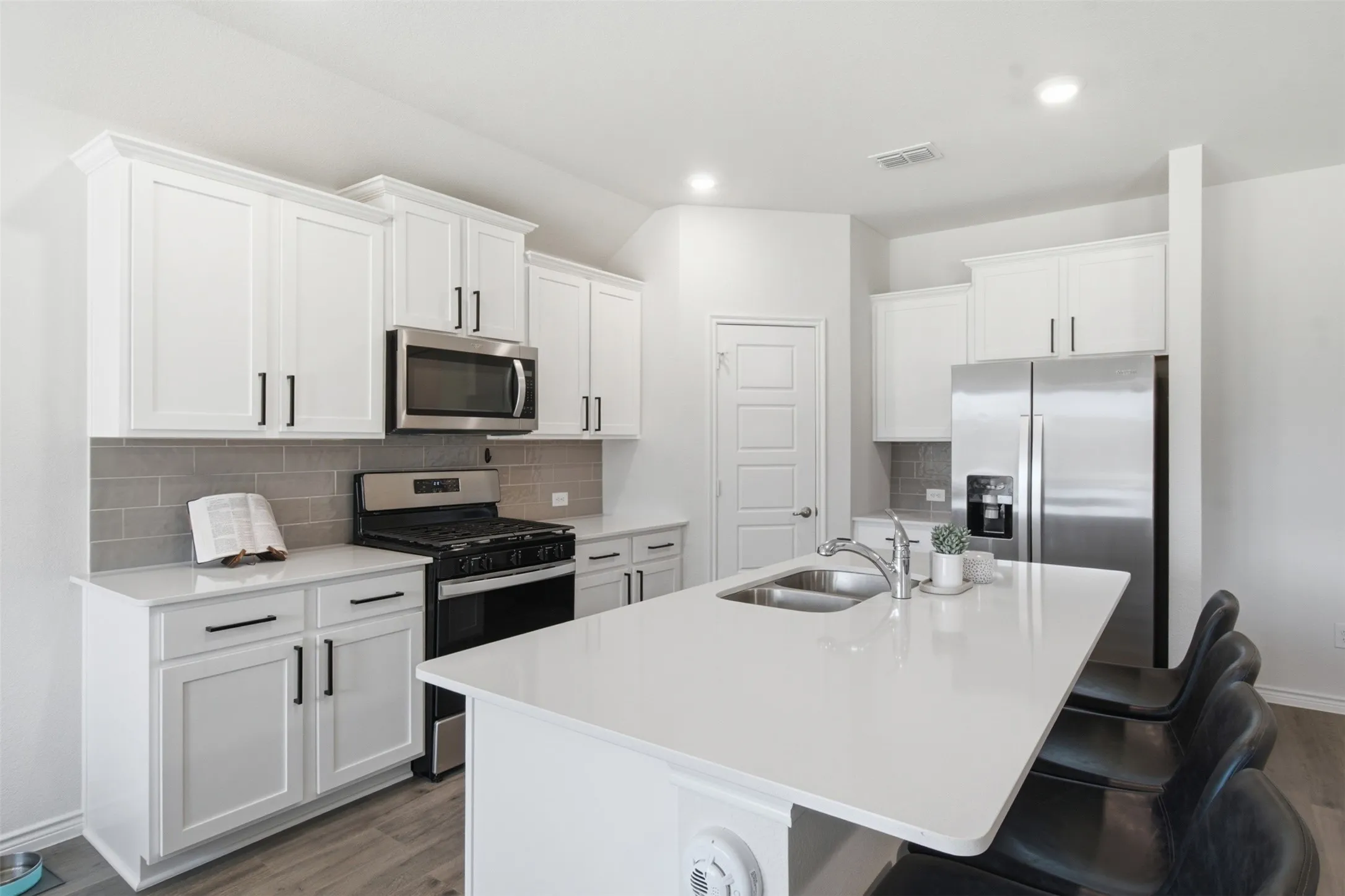 Kitchen featuring a central island with an integrated sink, white cabinetry, stainless steel appliances, a tiled backsplash, and wood-finish flooring