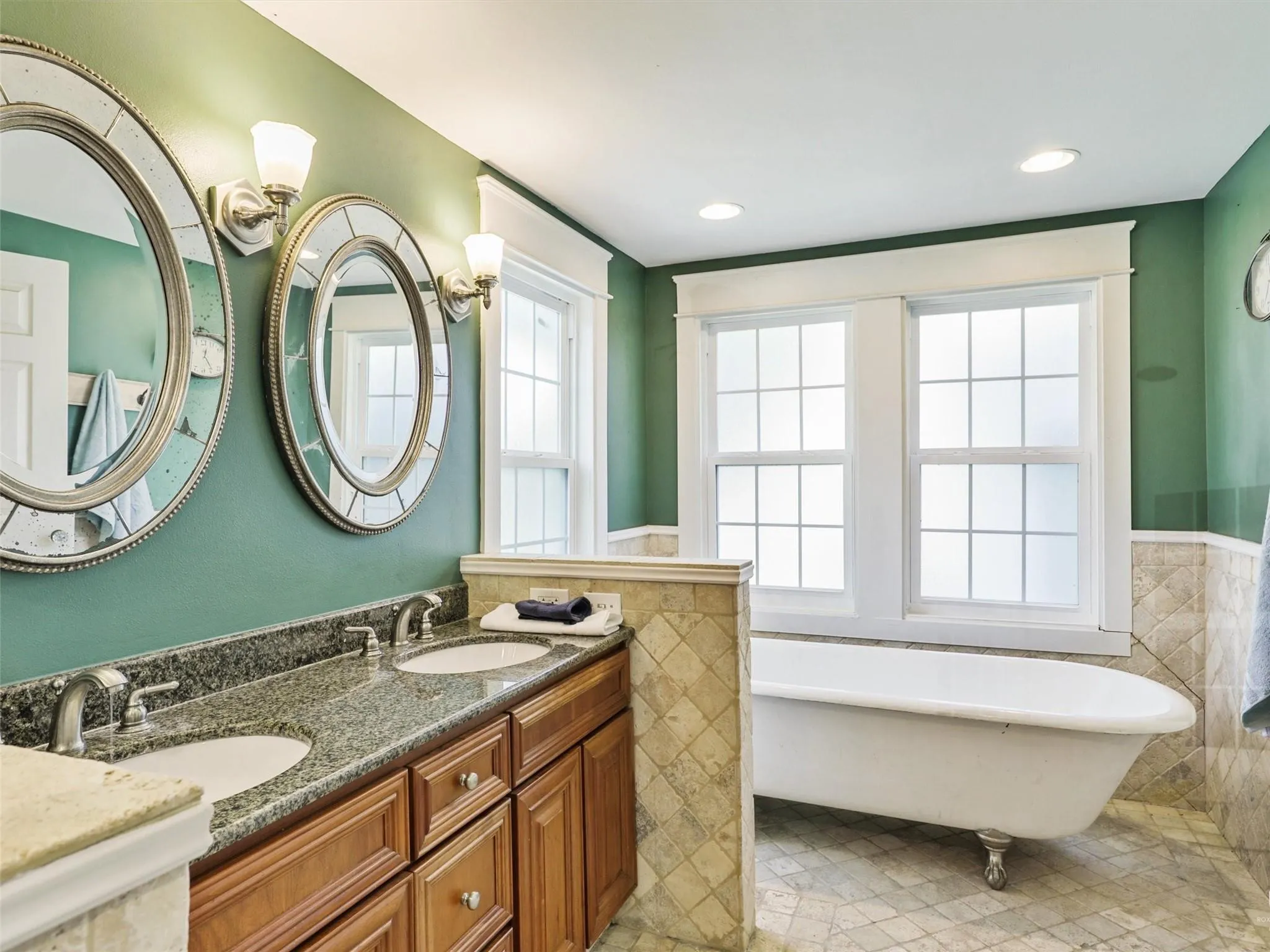 Bathroom featuring a clawfoot tub, dual windows, a double vanity with stone countertop, and wall-mounted sconces