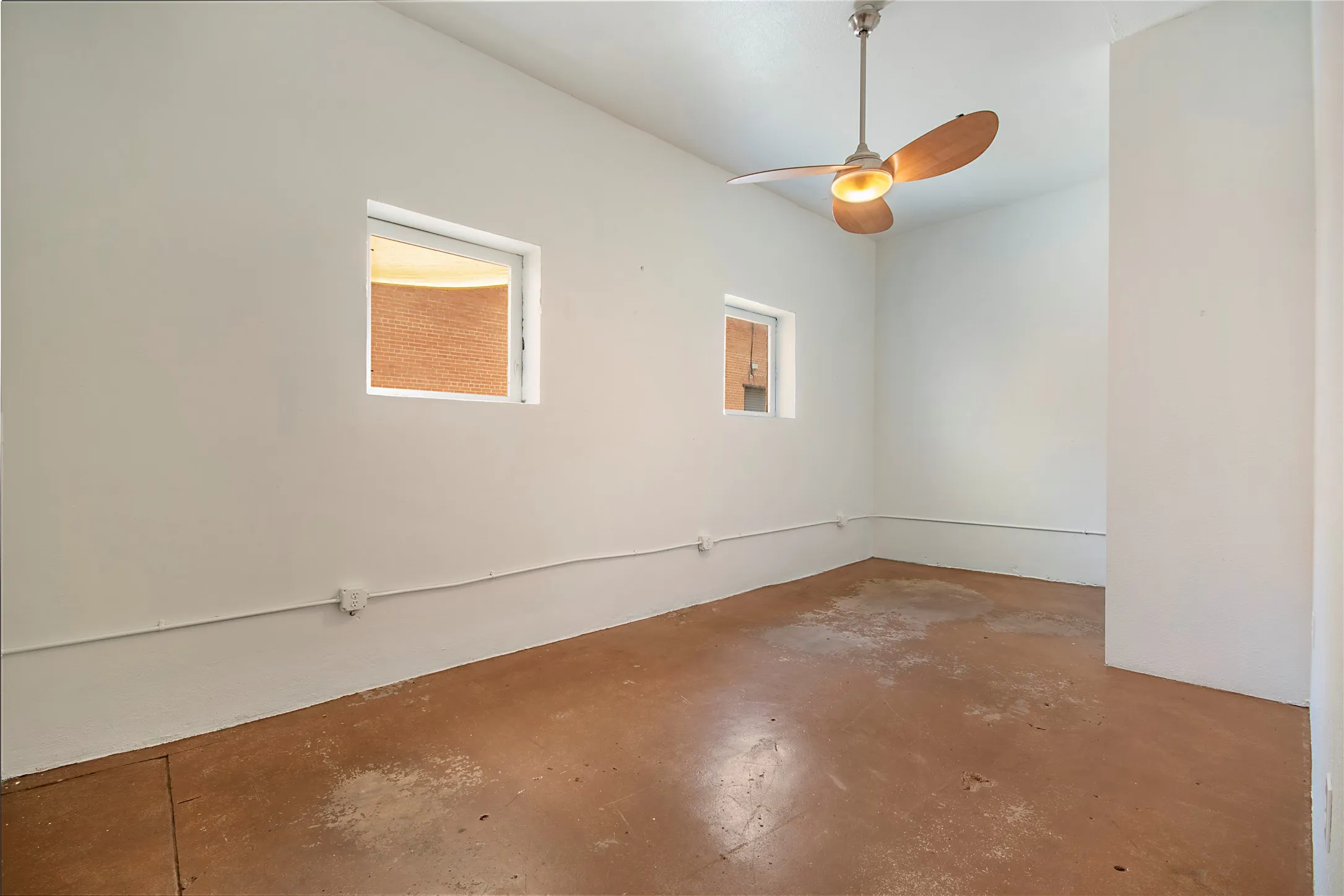 Room featuring white painted walls, a ceiling fan with light fixture, two windows, and earth-toned concrete flooring