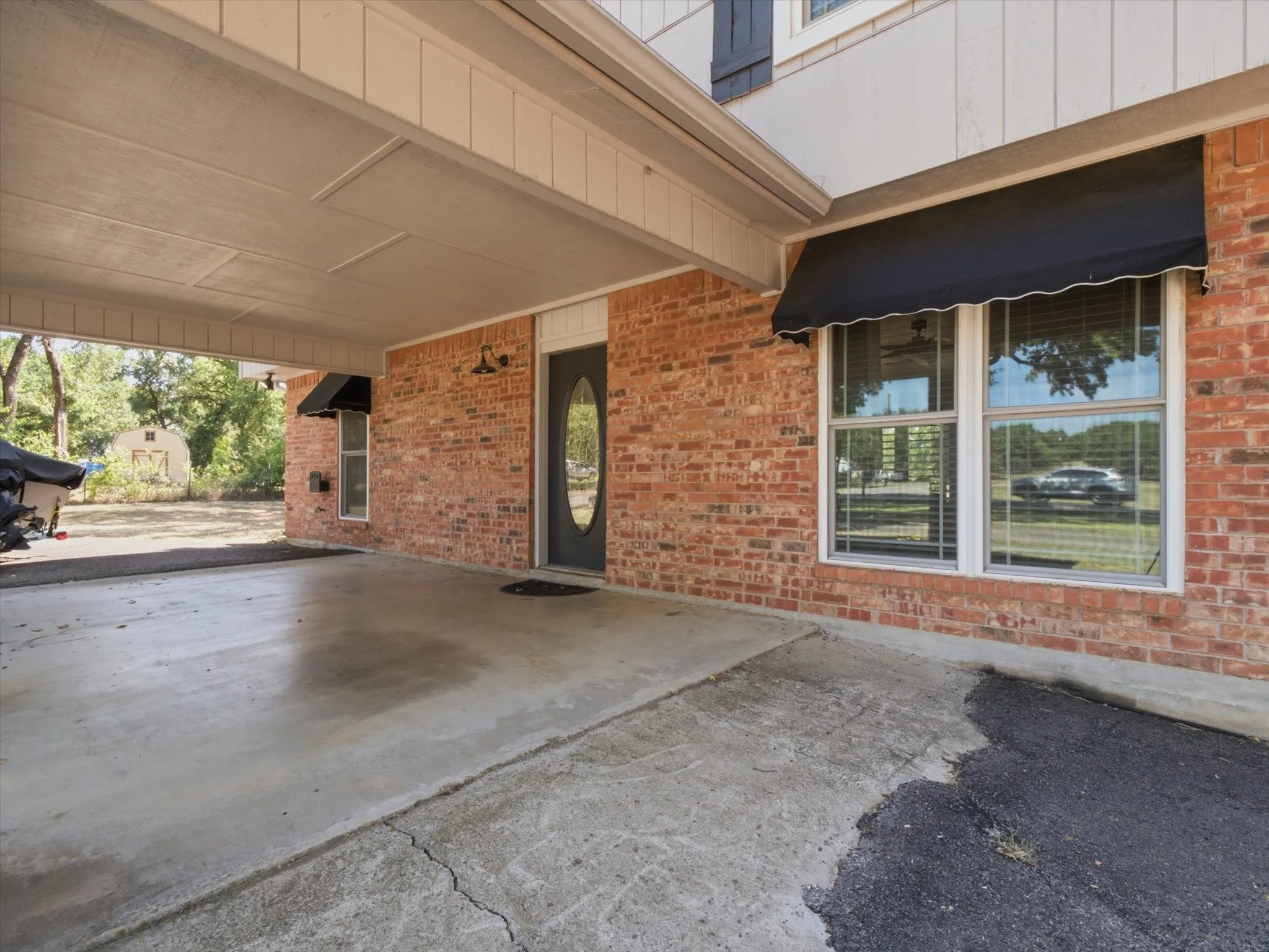 Brick facade entry with an oval glass front door, covered concrete carport, and a black window awning