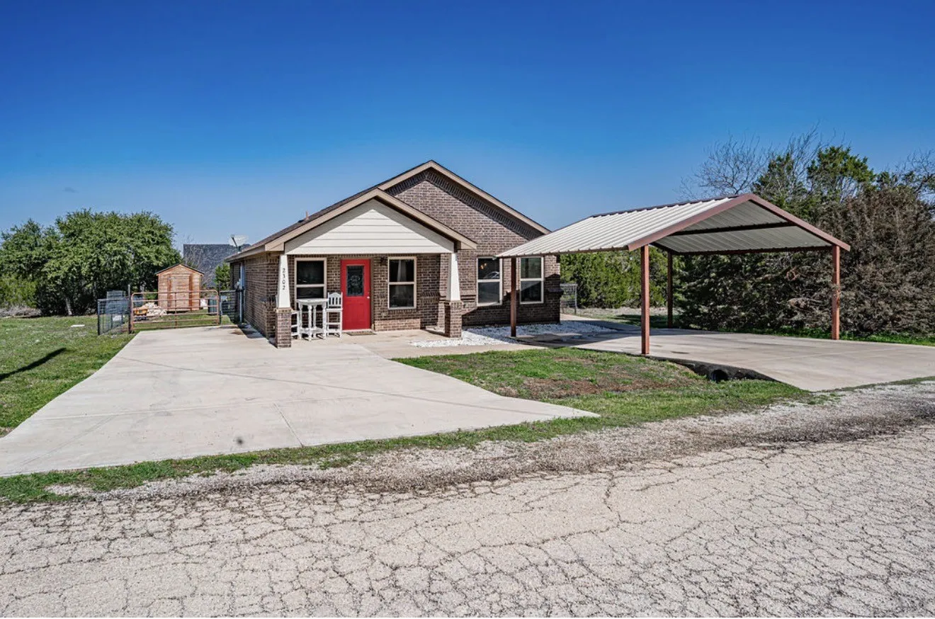 Brick exterior home featuring a covered front porch, red entry door, extended concrete driveway, and a detached carport