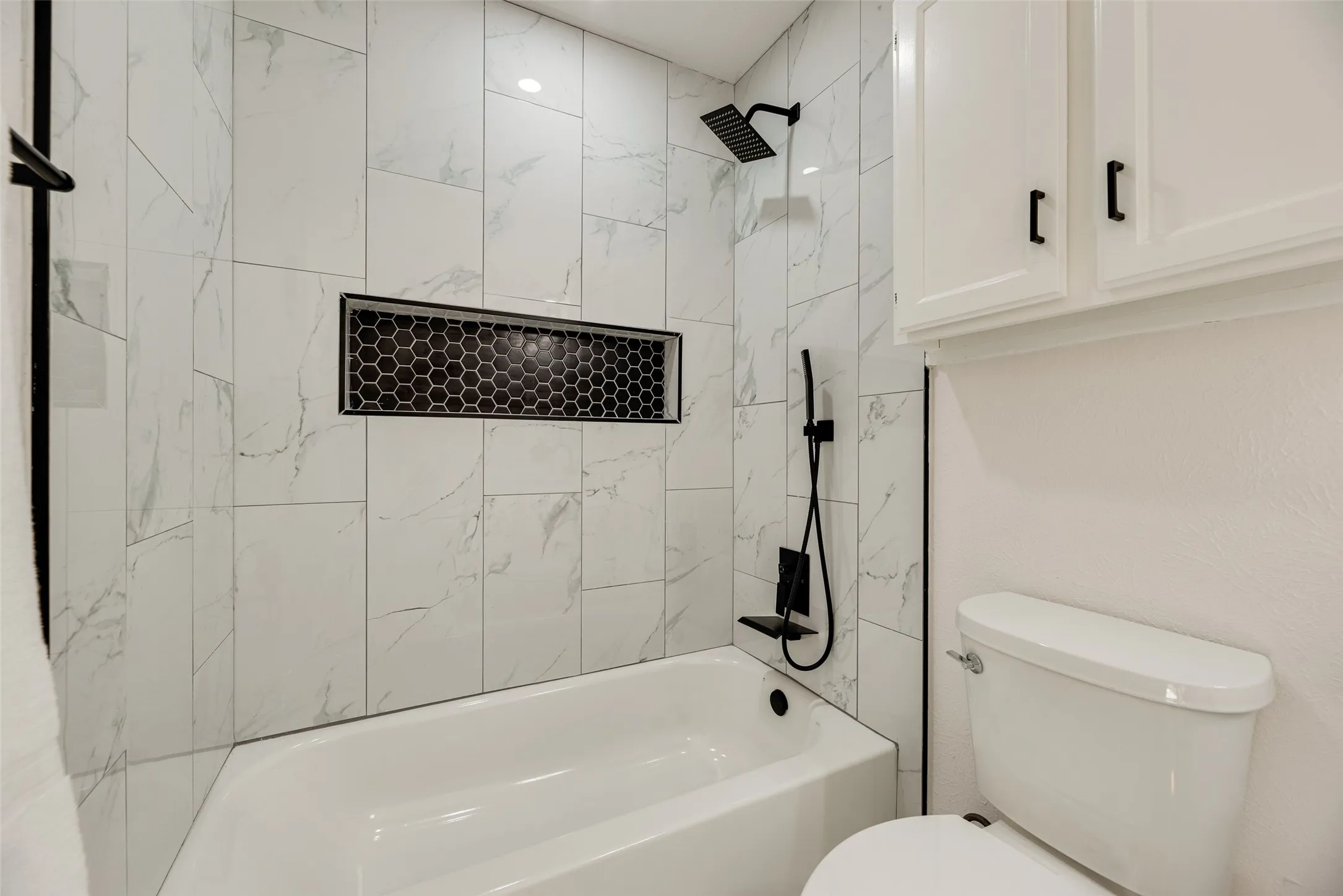 Bathtub and shower combination featuring large format white tile with grey veining, a black hexagonal tile shower niche, and matte black shower fixtures