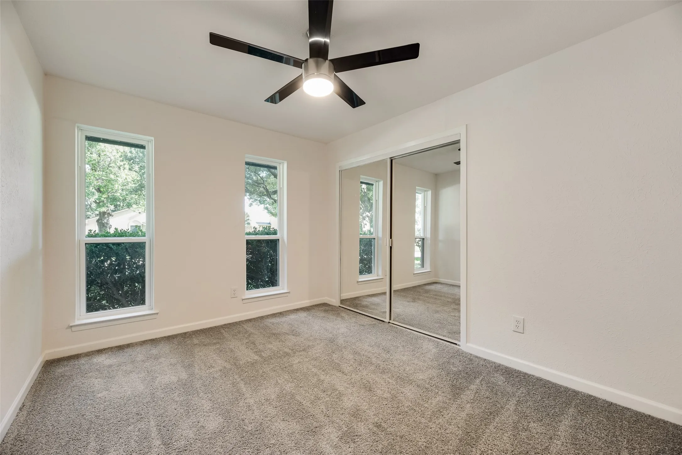 Carpeted room featuring two vertical windows with white trim, a contemporary ceiling fan, and mirrored closet doors