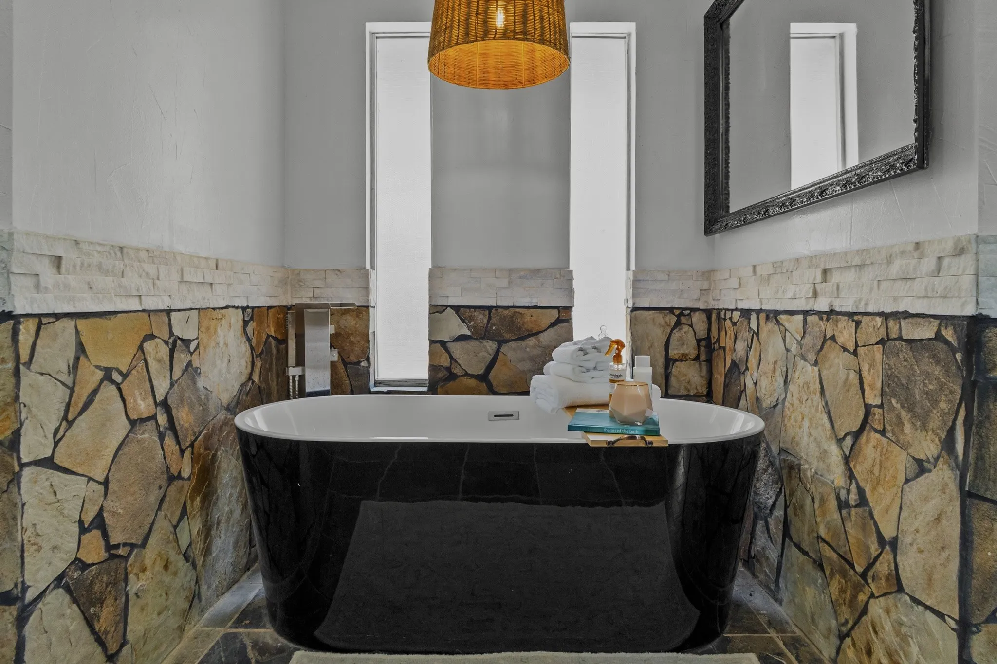 Freestanding black soaking tub with a stone-clad surround, complemented by a decorative mirror and woven pendant light fixture