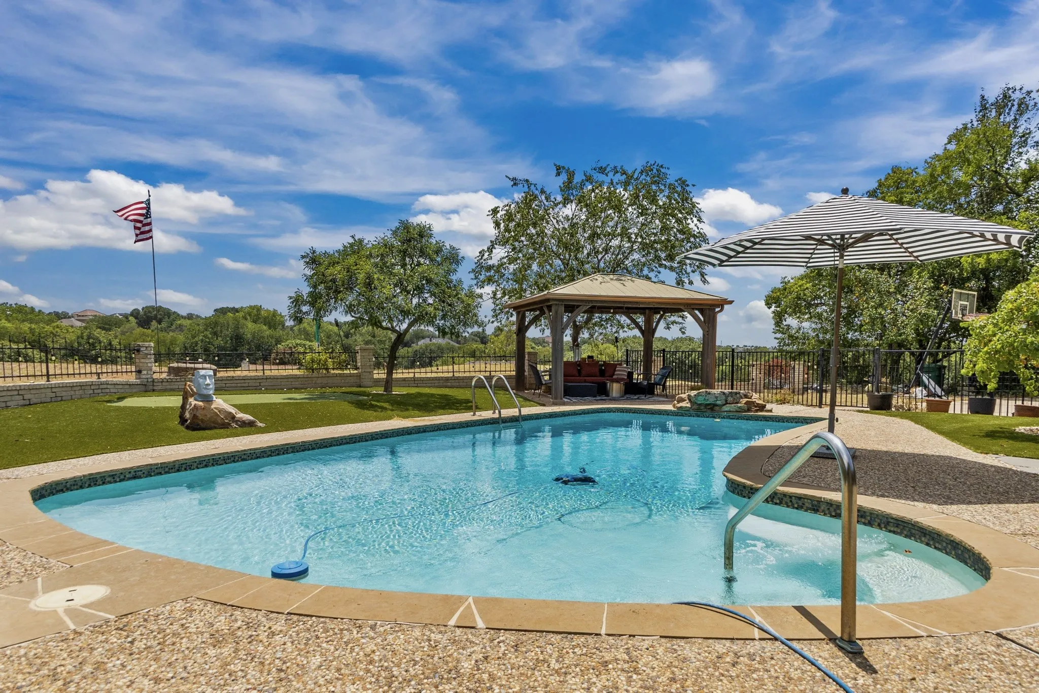 Outdoor pool with a surrounding aggregate concrete deck, a wooden gazebo, a putting green, and a wrought iron fence