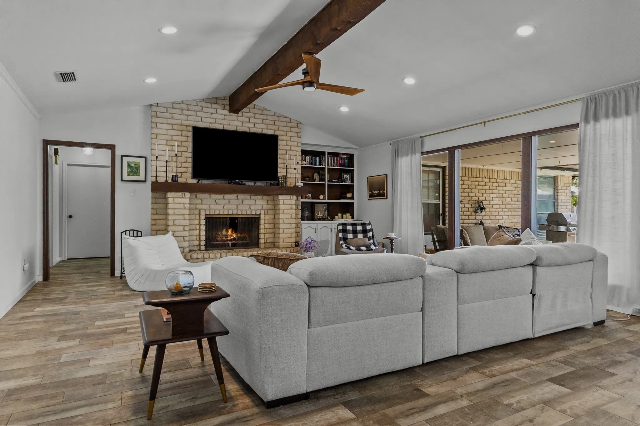 Vaulted ceiling with exposed wood beam, wood-finish flooring, brick fireplace with wood mantel, built-in shelving, and large sliding glass doors