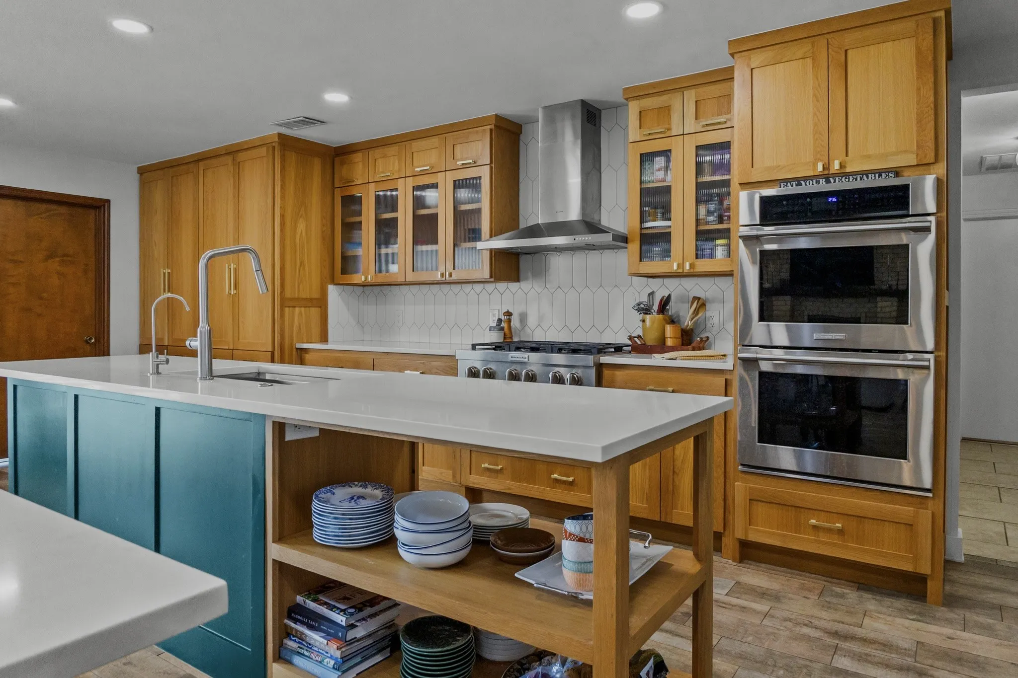 Kitchen featuring a central island with a white countertop and blue paneling, light wood cabinetry, stainless steel double wall ovens, a coordinating range hood, and a white tile backsplash