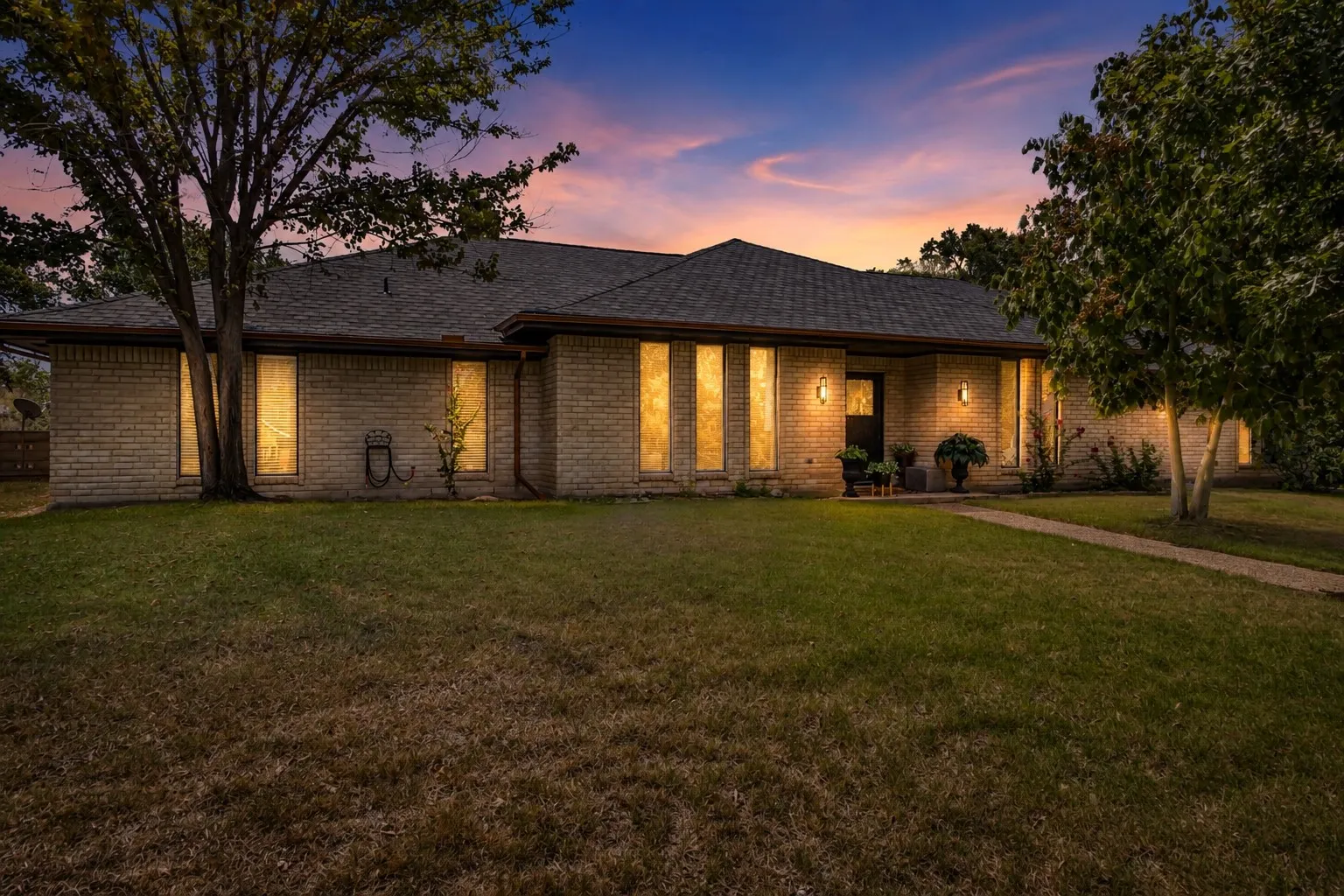 Brick exterior with a dark shingle roof, mature landscaping, and a concrete pathway